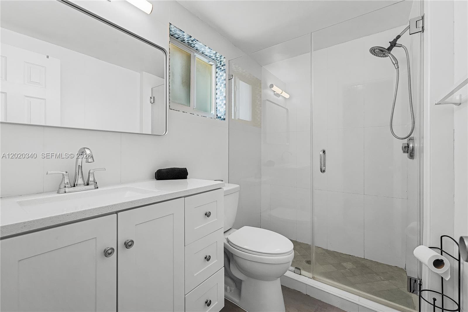A clean, all-white bathroom with a shaker-style vanity, quartz countertop, and large mirror. Features a glass-enclosed walk-in shower with a handheld showerhead and a pop of blue mosaic tile framing the frosted window. Simple and fresh.
