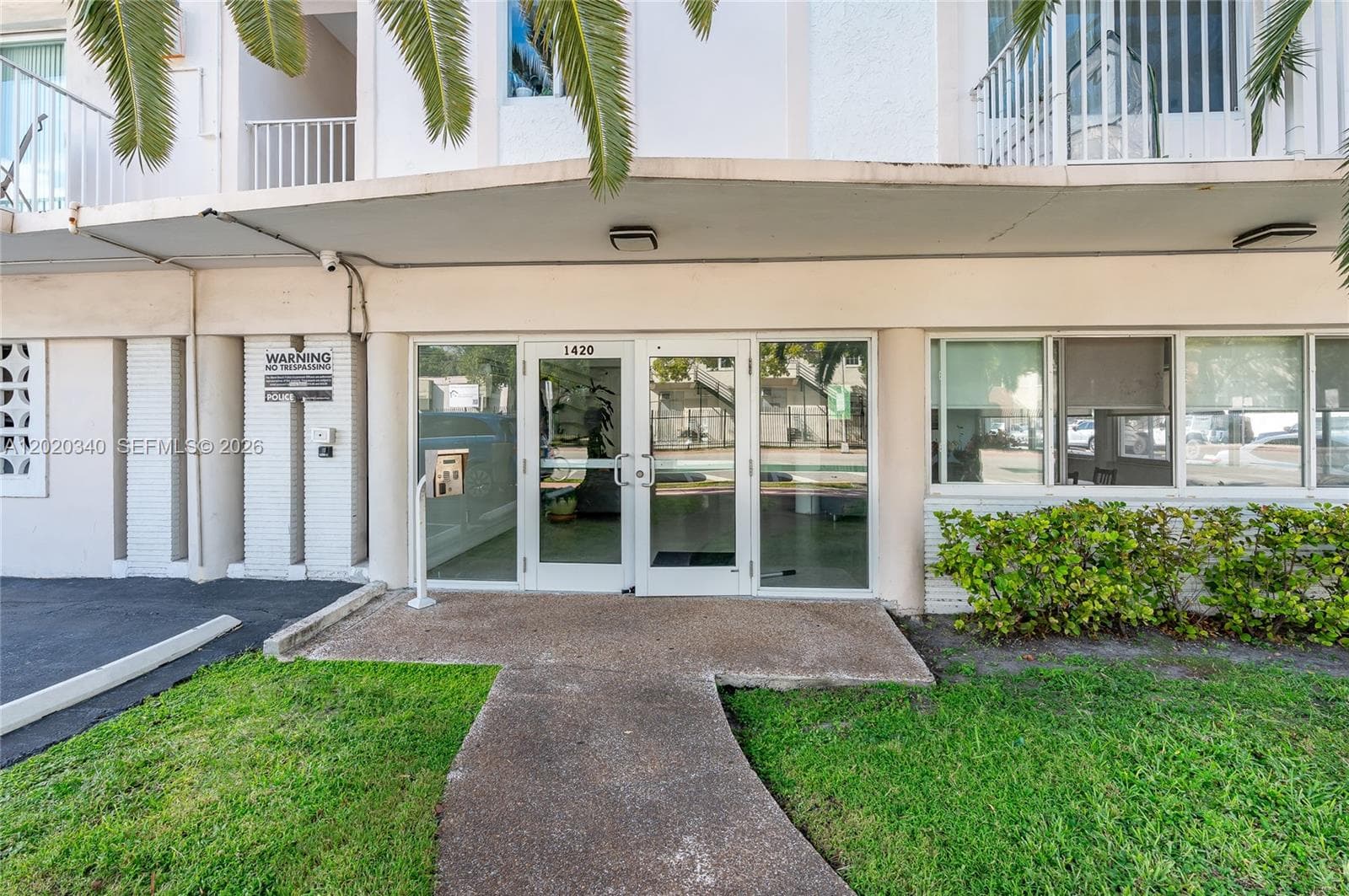 The ground-floor entrance of a mid-rise residential building at 1420, featuring glass double doors with a secured intercom system. The exterior is finished in cream-toned stucco with white-framed balconies visible above, framed by tropical palm trees and neatly trimmed hedges. A concrete walkway leads from the street to the lobby entrance, with a glimpse of a pool courtyard through the glass doors.