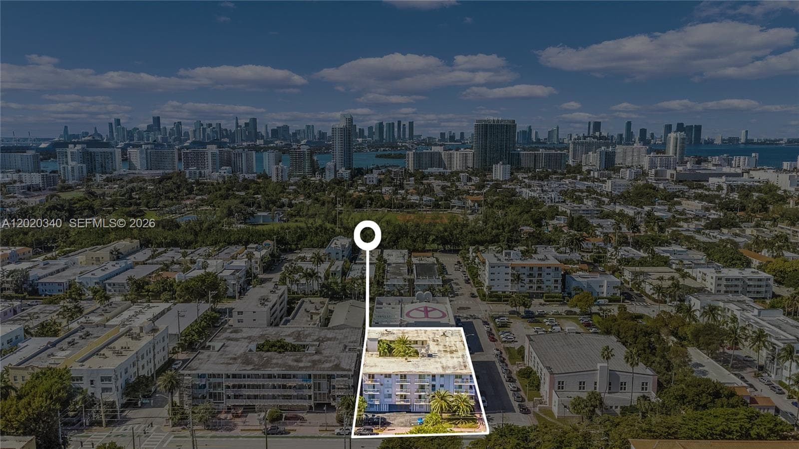 Aerial view showing the building's prime Miami Beach location — a low-rise mid-century building nestled in a lush, tree-lined neighborhood with the downtown Miami skyline and Biscayne Bay visible in the background.