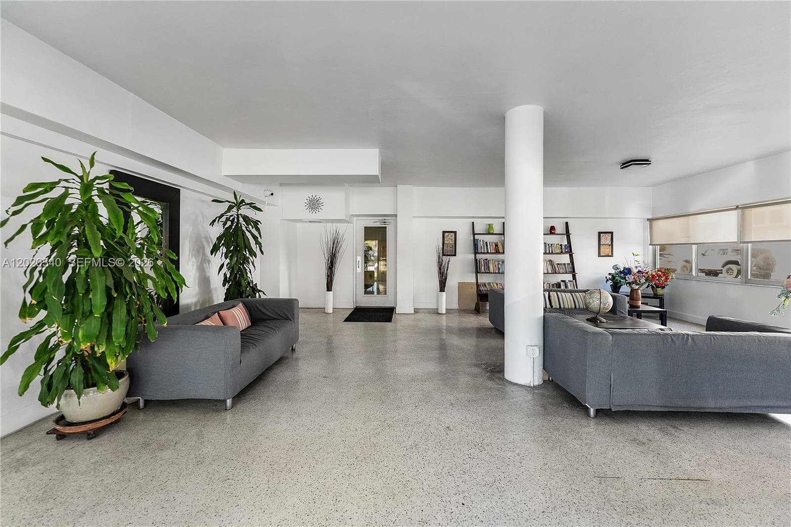 A spacious, loft-style common or living area with polished concrete floors, twin gray sofas, and a prominent white column. Large tropical plants, ladder bookshelves, and dried branch arrangements give it an airy, urban-jungle feel.