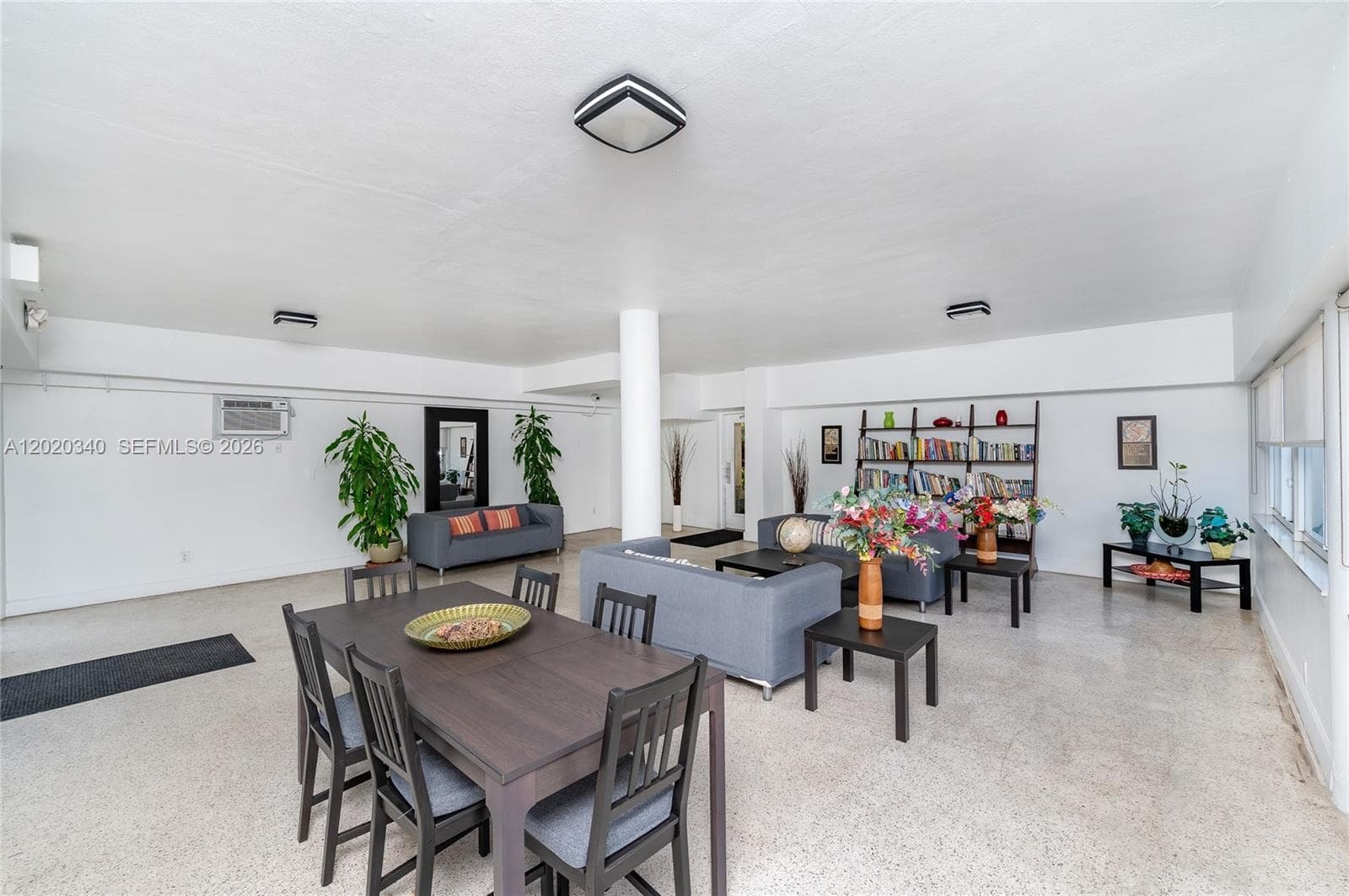 A spacious building common room with terrazzo floors, a dining table, gray sofas, ladder bookshelf, and tropical plants — relaxed and social, with plenty of room to gather.