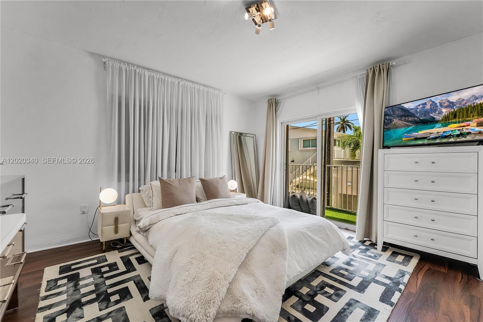 A bright bedroom with a plush king-size bed, globe bedside lamps, and a bold black-and-white geometric rug on dark hardwood floors. A sliding glass door opens to the balcony, flanked by neutral curtains, with a white dresser and wall-mounted TV to the side.