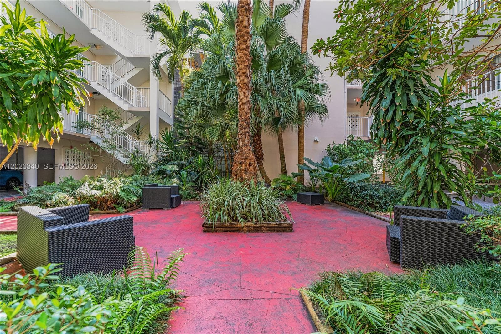A lush tropical courtyard at the center of a multi-story building, with tall palm trees, dense greenery, and black wicker seating on a bold red-painted patio. Private, garden-like, and very South Florida.
