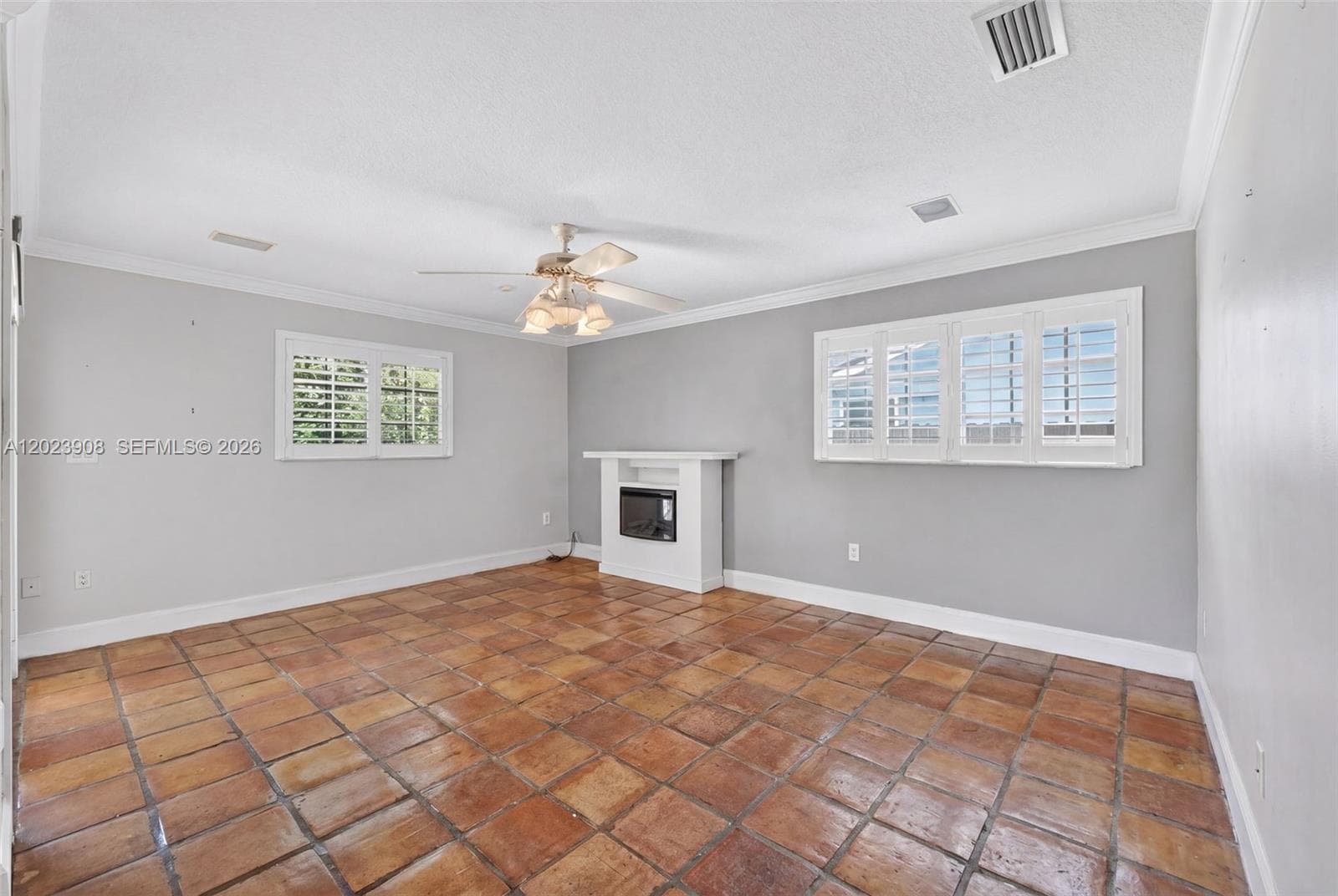 Living area featuring terracotta tile flooring, crown molding, plantation shutters, and a built-in electric fireplace with a mantel