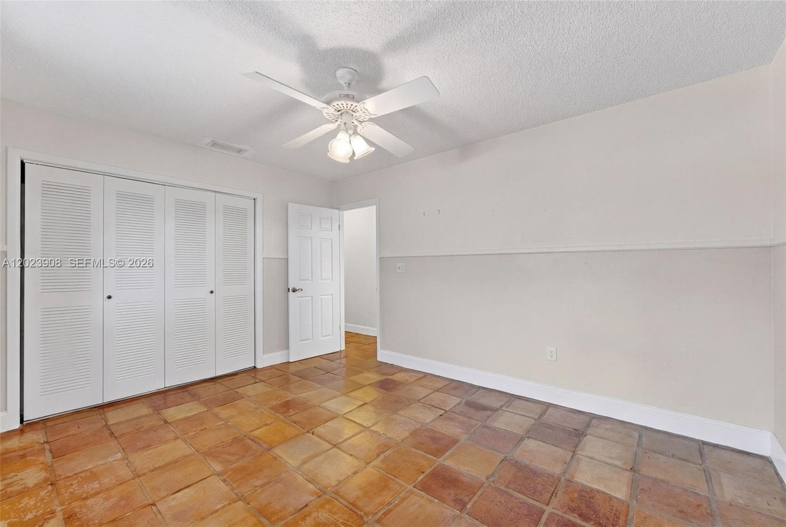 Room featuring square terracotta-style tile flooring, dual louvered-panel closet doors, a multi-panel interior door, and a ceiling-mounted fan with integrated light fixtures