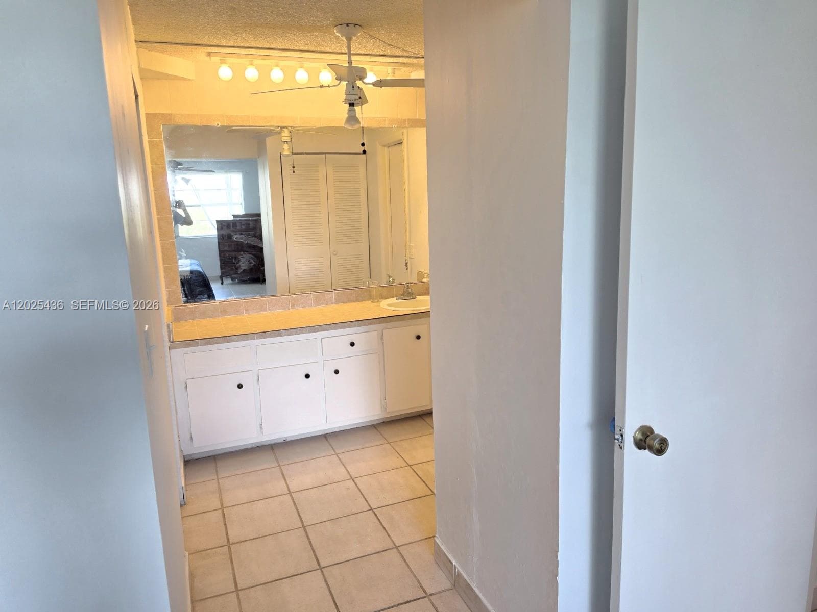 Bathroom featuring a white vanity cabinet, solid-surface countertop with an integrated sink, expansive wall-mounted mirror, ceramic tile flooring, and ceiling fan with integrated lighting
