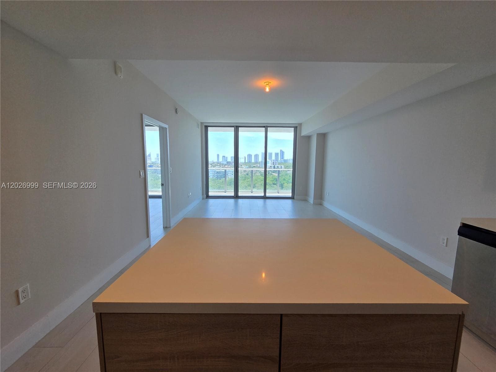 Open-concept living area flows into a modern kitchen with a central island, light wood cabinetry, and stainless steel appliances. The neutral finishes and continuous tile flooring create a clean, cohesive layout.