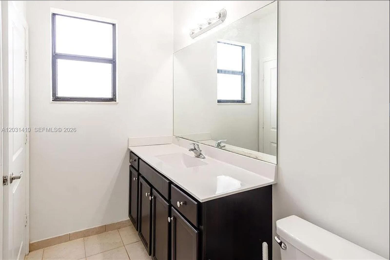 Vanity featuring dark-finish cabinetry, a white countertop with an integrated sink, a large wall-mounted mirror, and tile flooring