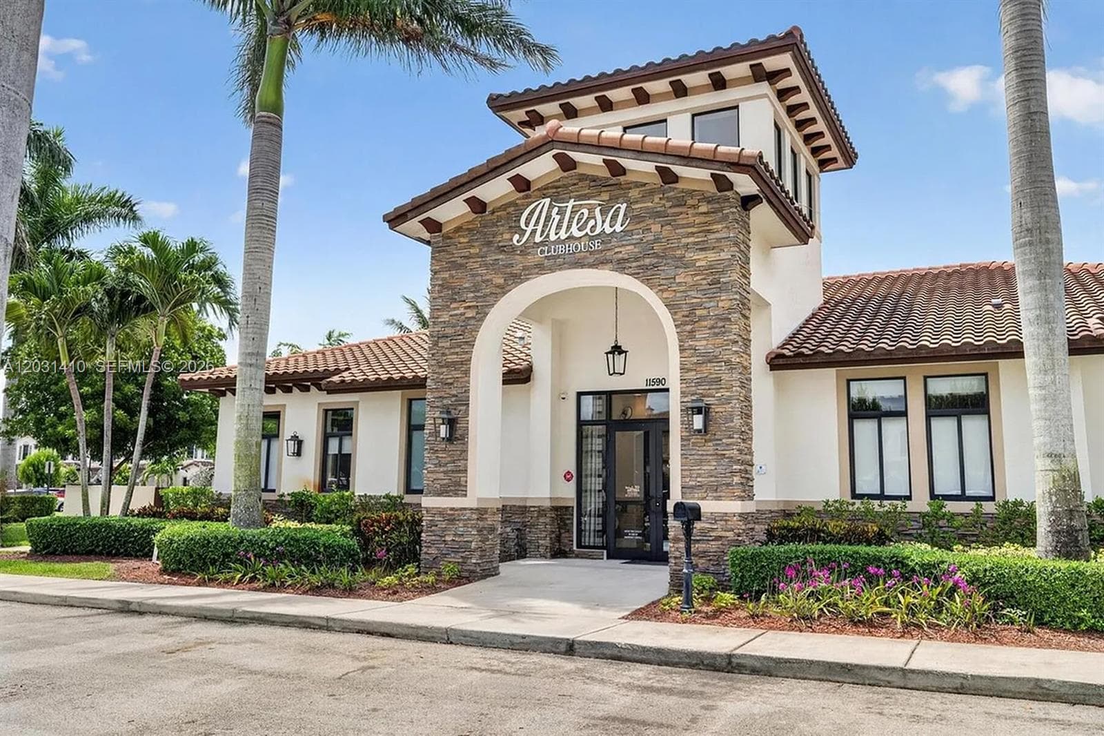 Clubhouse exterior featuring stacked stone masonry facade, arched entryway, clay tile roofing, decorative wood corbels, and manicured perimeter landscaping