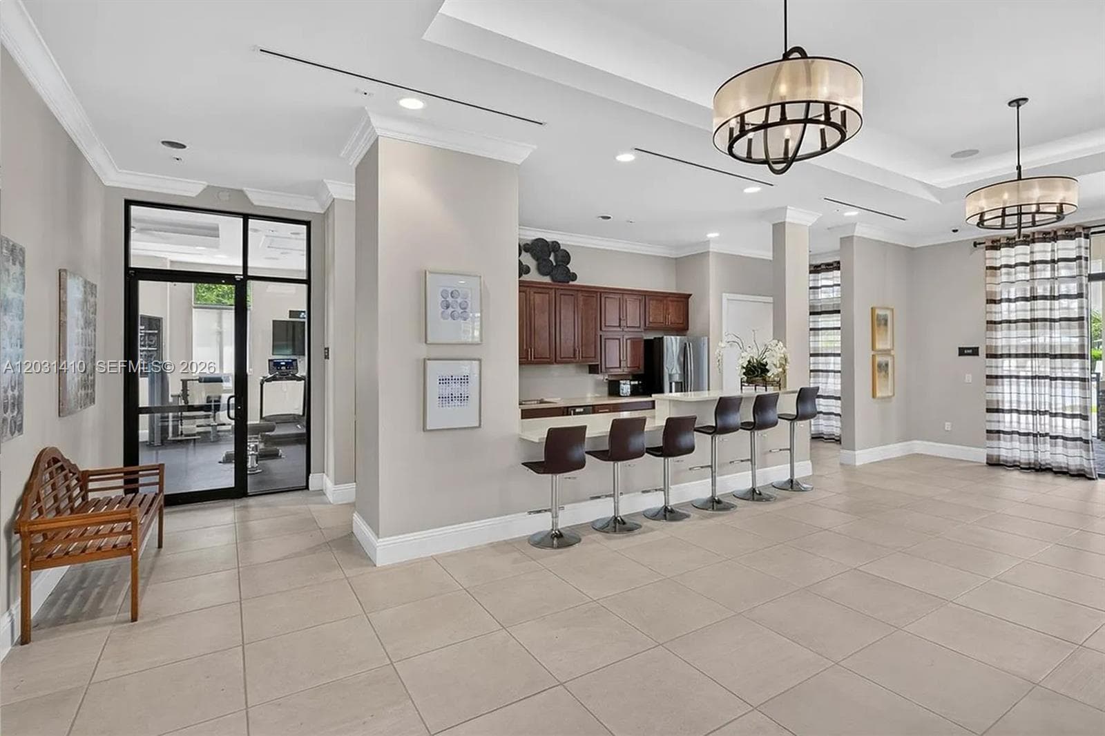 Open-concept common area featuring large-format tile flooring, tray ceilings with recessed lighting, two drum-style chandeliers, a built-in refreshment bar with cabinetry, and a glass-paneled entry door
