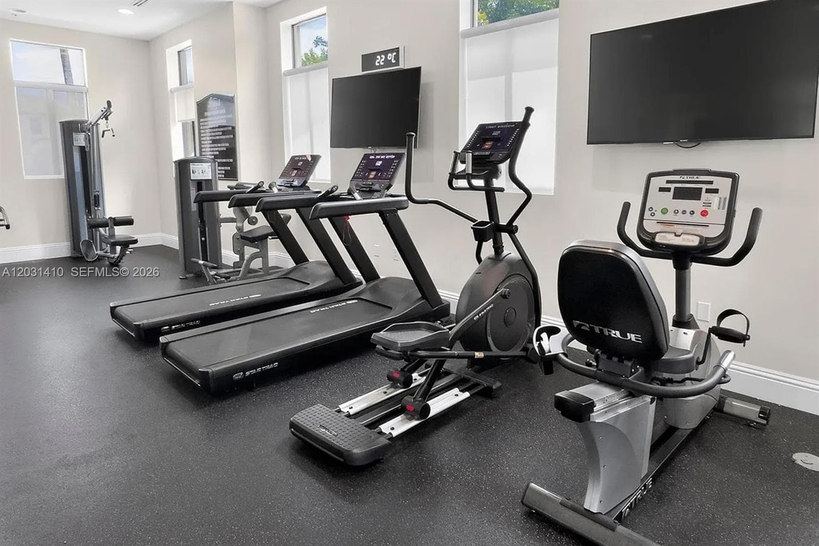 Fitness center featuring wall-mounted televisions, large windows with neutral roller shades, and impact-resistant rubberized flooring