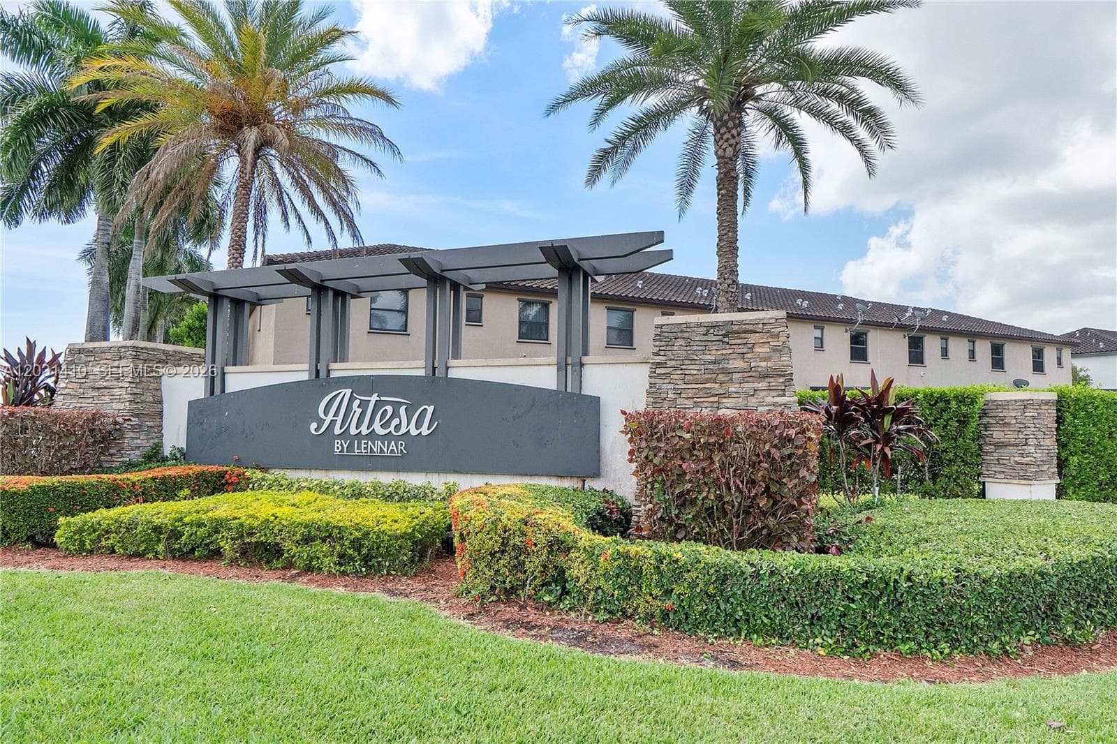 Community entrance featuring a monument sign, stacked stone pillars, decorative metal pergola, mature palm trees, and manicured hedges