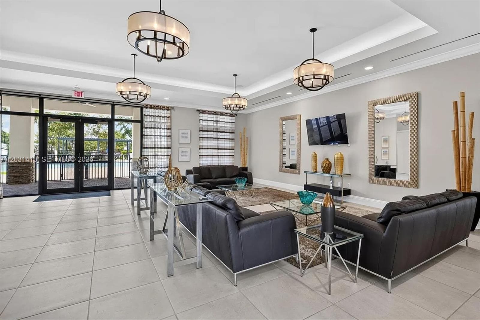 Spacious room featuring neutral tile flooring, recessed tray ceiling, multiple tiered drum-style pendant light fixtures, and expansive glass-paneled double doors