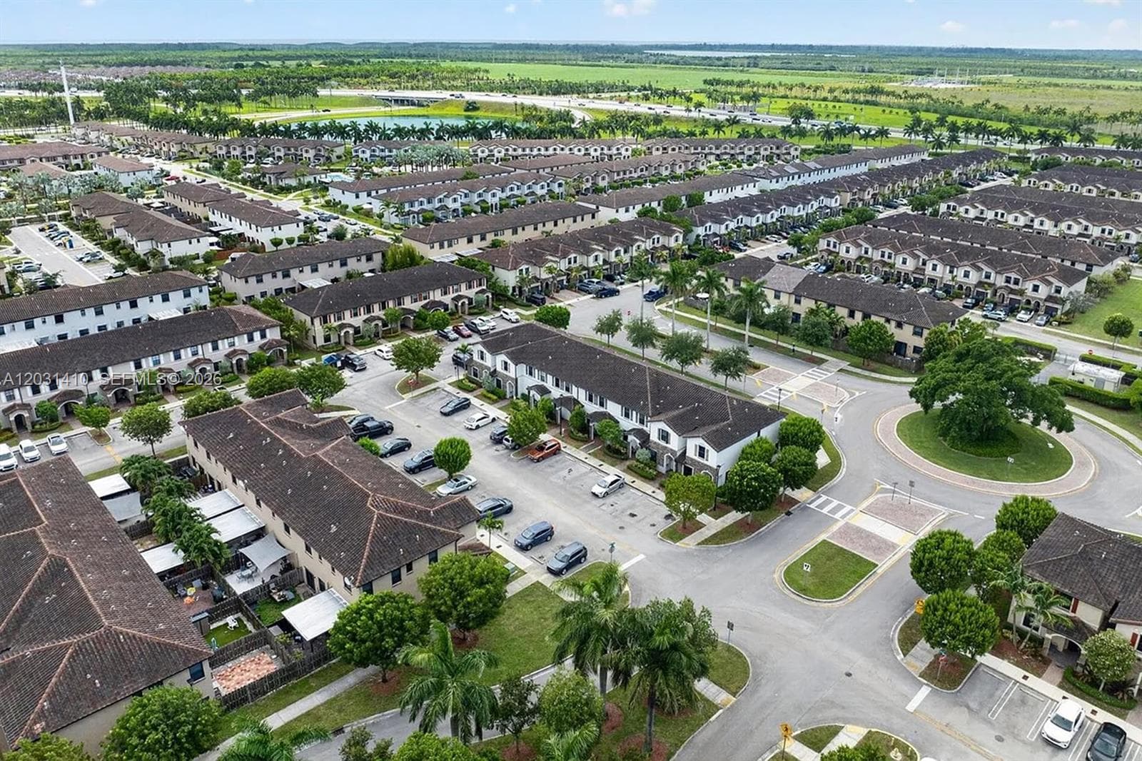 Aerial view of residential community featuring multi-unit structures with dark-toned shingle roofing, paved roadways, designated surface parking, and manicured landscaping including mature palm trees