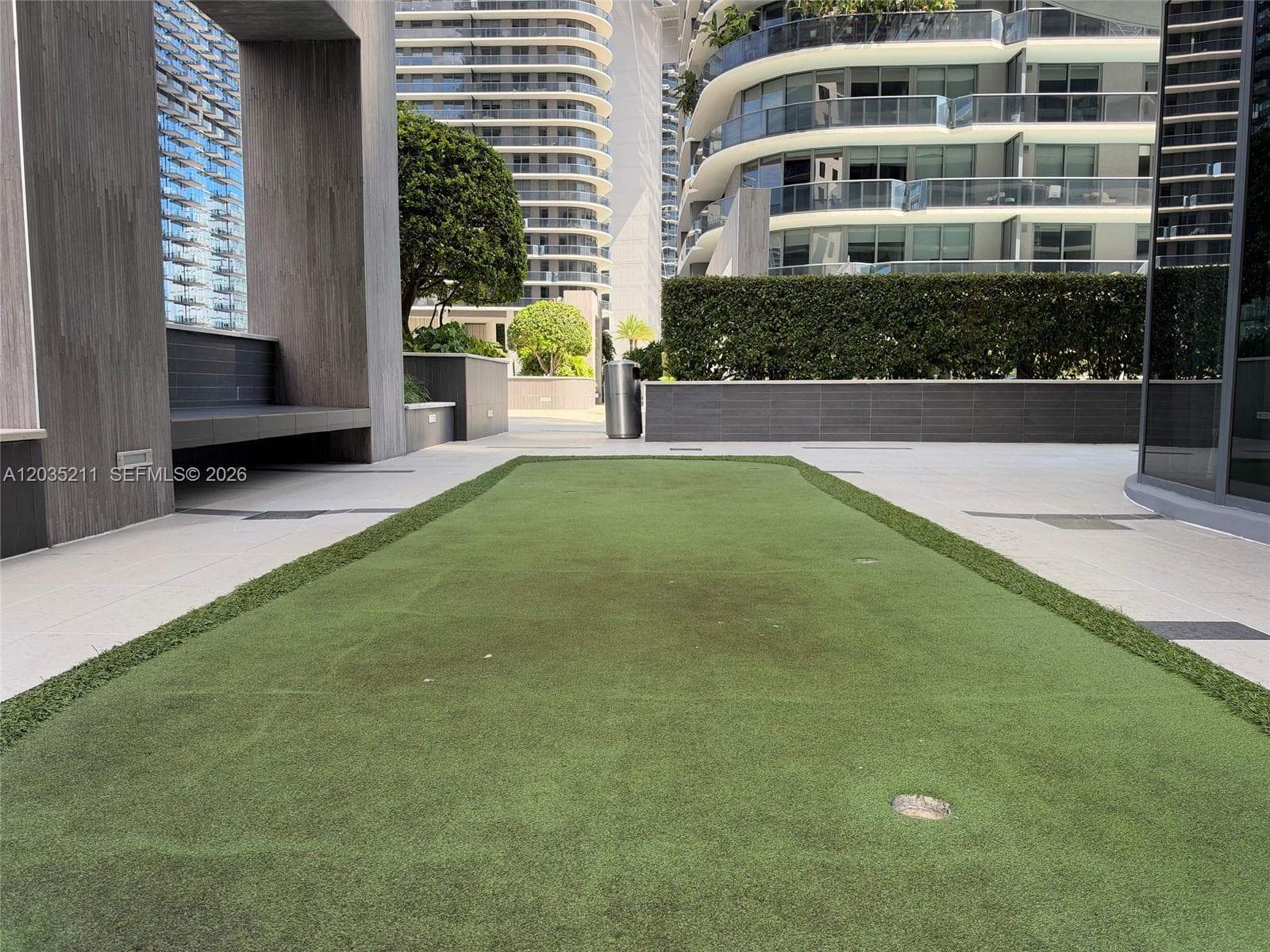 Outdoor putting green with synthetic turf, concrete terrace flooring, integrated stone-finish bench seating, and manicured hedge perimeter