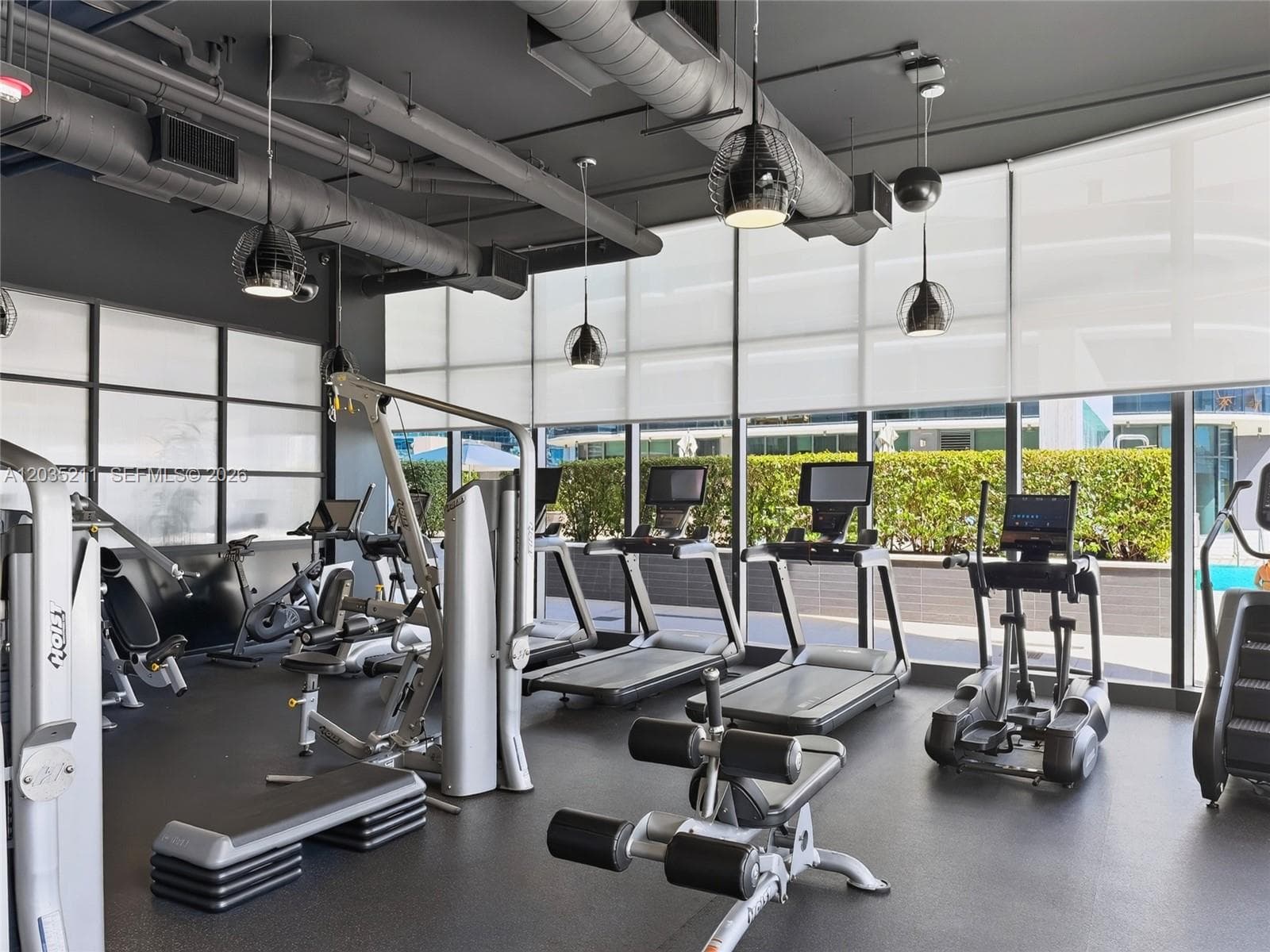 Fitness space featuring floor-to-ceiling windows with roller shades, industrial-style exposed ductwork, and dark rubberized flooring