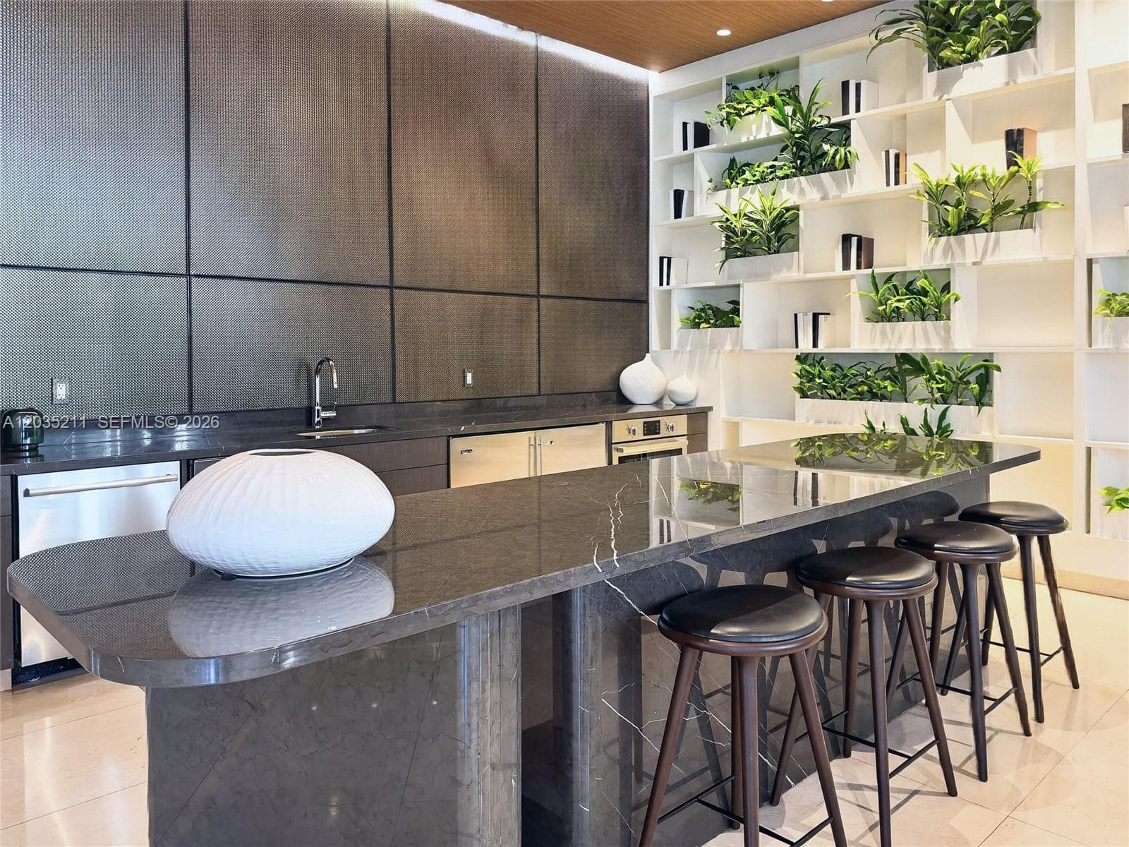 Kitchen featuring a stone-topped island with waterfall edge, full-height decorative panel wall, built-in floor-to-ceiling shelving, stainless steel appliances, and integrated under-cabinet lighting