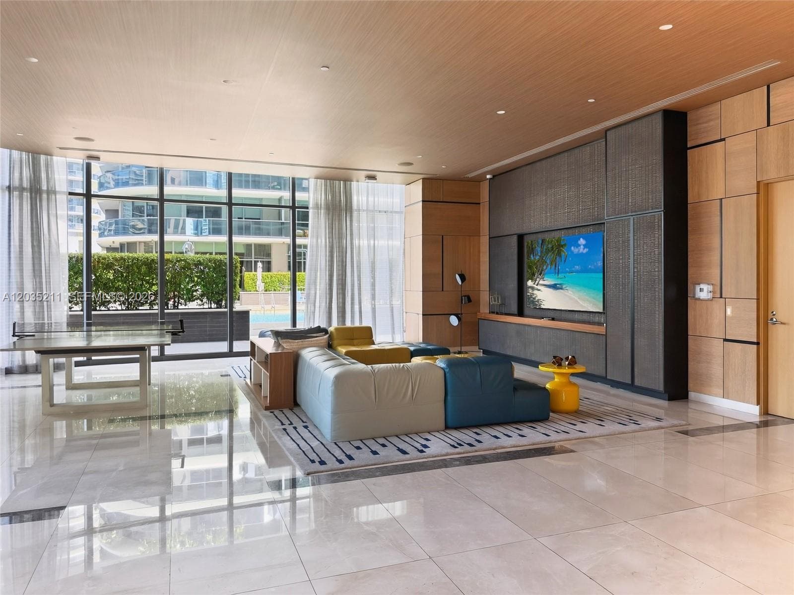 Floor-to-ceiling glass wall, light wood paneling, large-format tile flooring, recessed ceiling lighting, and built-in media cabinetry with metallic finish