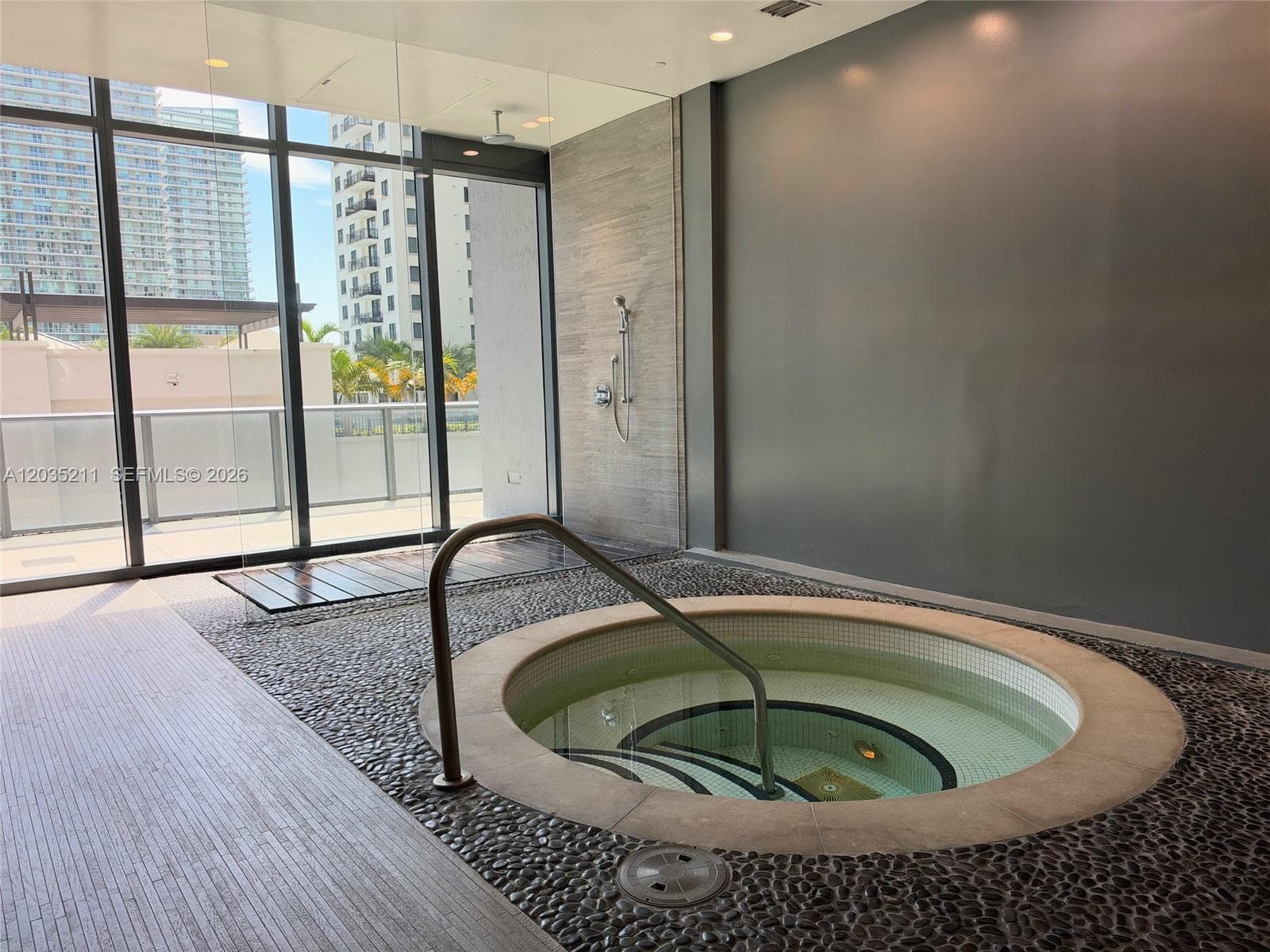 In-ground circular spa featuring a pebble-stone surround, metal handrail, full-height floor-to-ceiling windows, and wall-mounted shower fixture