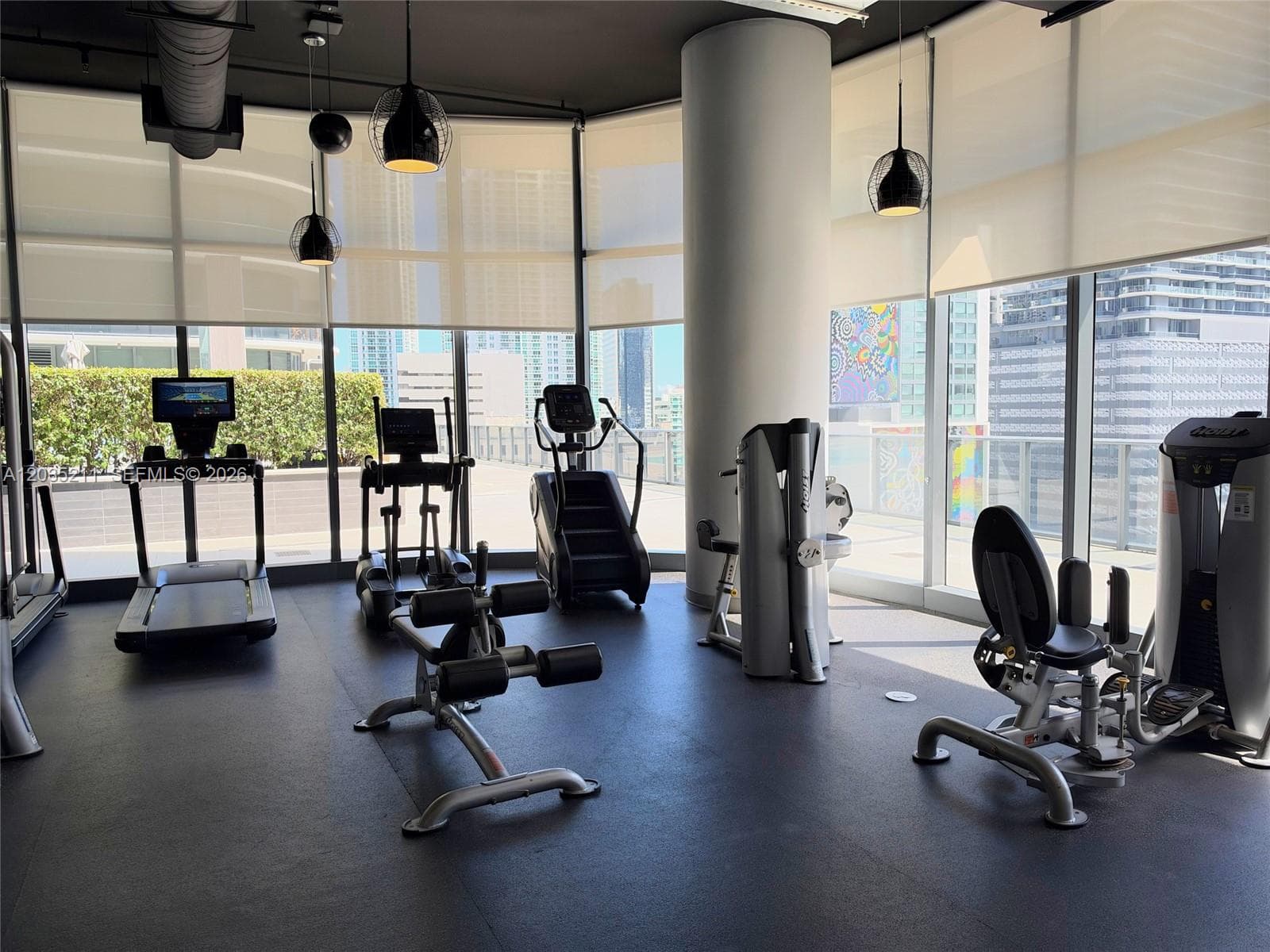 Fitness center featuring floor-to-ceiling windows, rubberized flooring, exposed ductwork, pendant lighting, and automated roller shades
