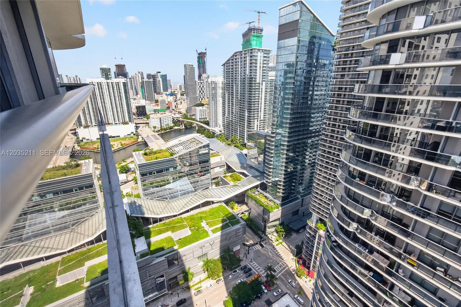 Elevated balcony featuring a metal railing with a clear glass insert, overlooking an urban cityscape with high-rise structures and green-roof architecture