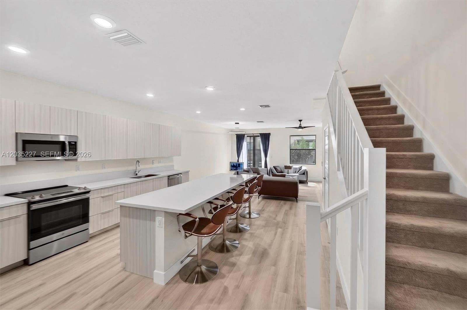 Bright open-concept floor plan connecting the kitchen, dining island, and living area in seamless flow. Sleek stainless appliances and a long quartz island with bar seating set the scene, with a carpeted staircase adding architectural interest.