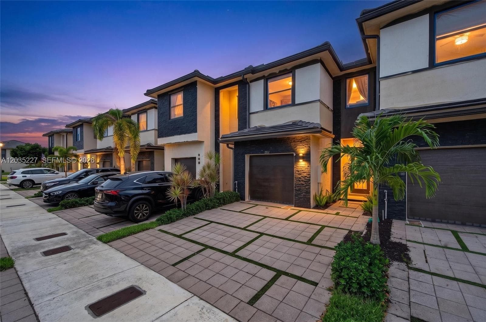 Captivating twilight street view of a contemporary townhome community, featuring dramatic stacked-stone facades and warm illuminated windows. Mature palm trees and decorative paver driveways enhance the upscale curb appeal under a stunning sunset sky.