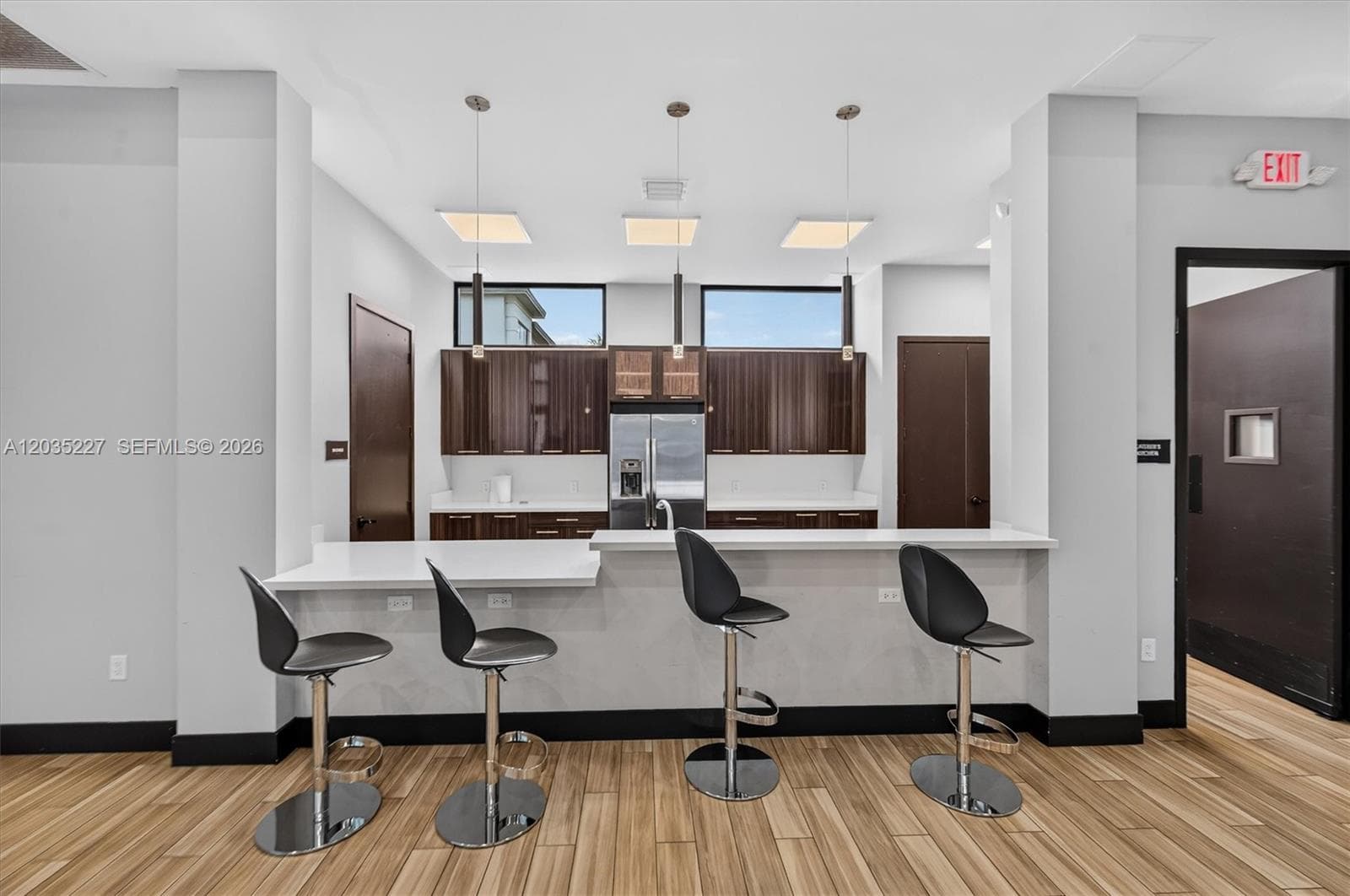 Trendy community kitchen lounge featuring rich espresso cabinetry and a stylish bar with sleek black stools. Pendant lighting and clerestory windows add a chic, sophisticated touch above the warm wood flooring.