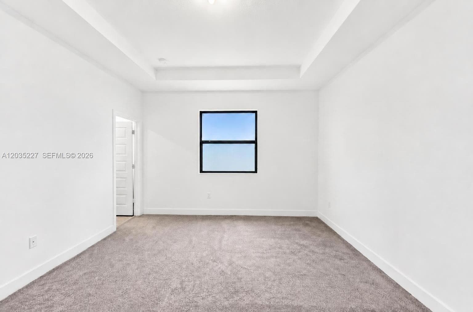 Spacious bedroom with plush carpeting and a tray ceiling that adds architectural detail. A large window brings in natural light, complemented by a cordless shade for privacy. Neutral tones throughout offer a blank canvas for personal style.
