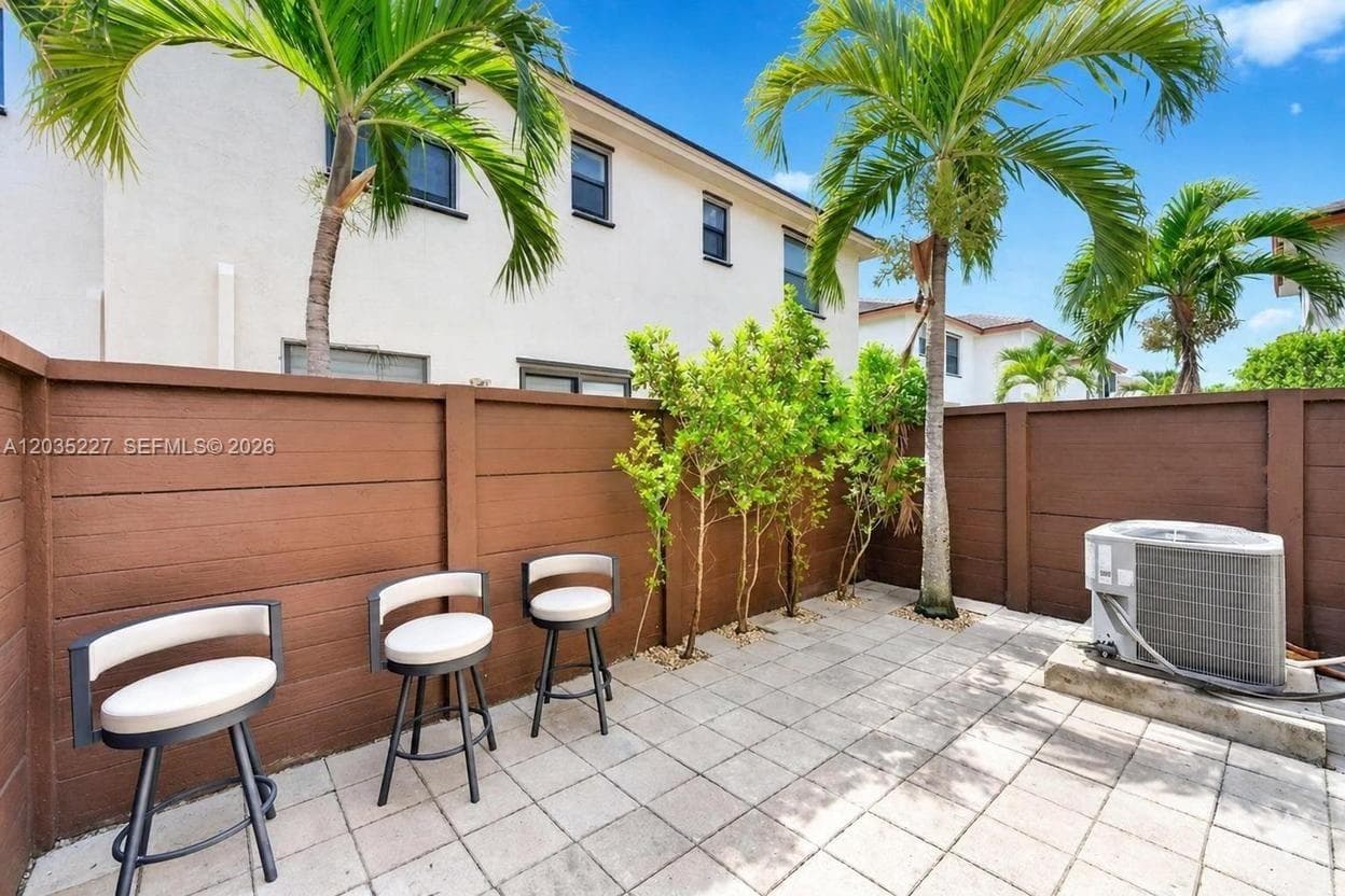 Sun-drenched private courtyard featuring towering palm trees and a wood privacy fence for added seclusion. Paver flooring and casual seating make this charming outdoor space ideal for relaxing or entertaining.