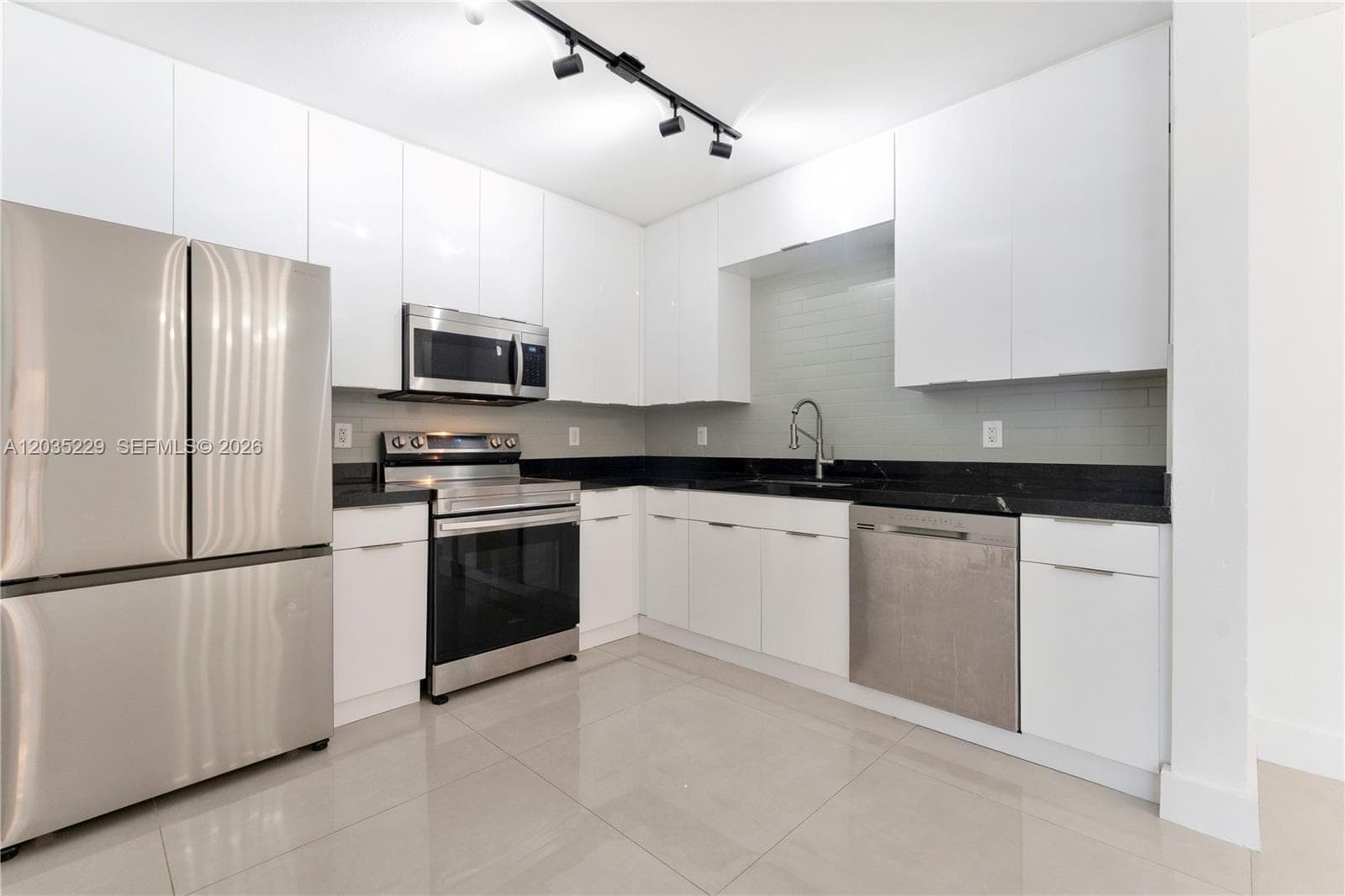 Angled view showcasing the full kitchen layout — floor-to-ceiling white cabinetry, dark granite countertops, full stainless steel appliance suite, and gray subway tile backsplash with polished tile floors.