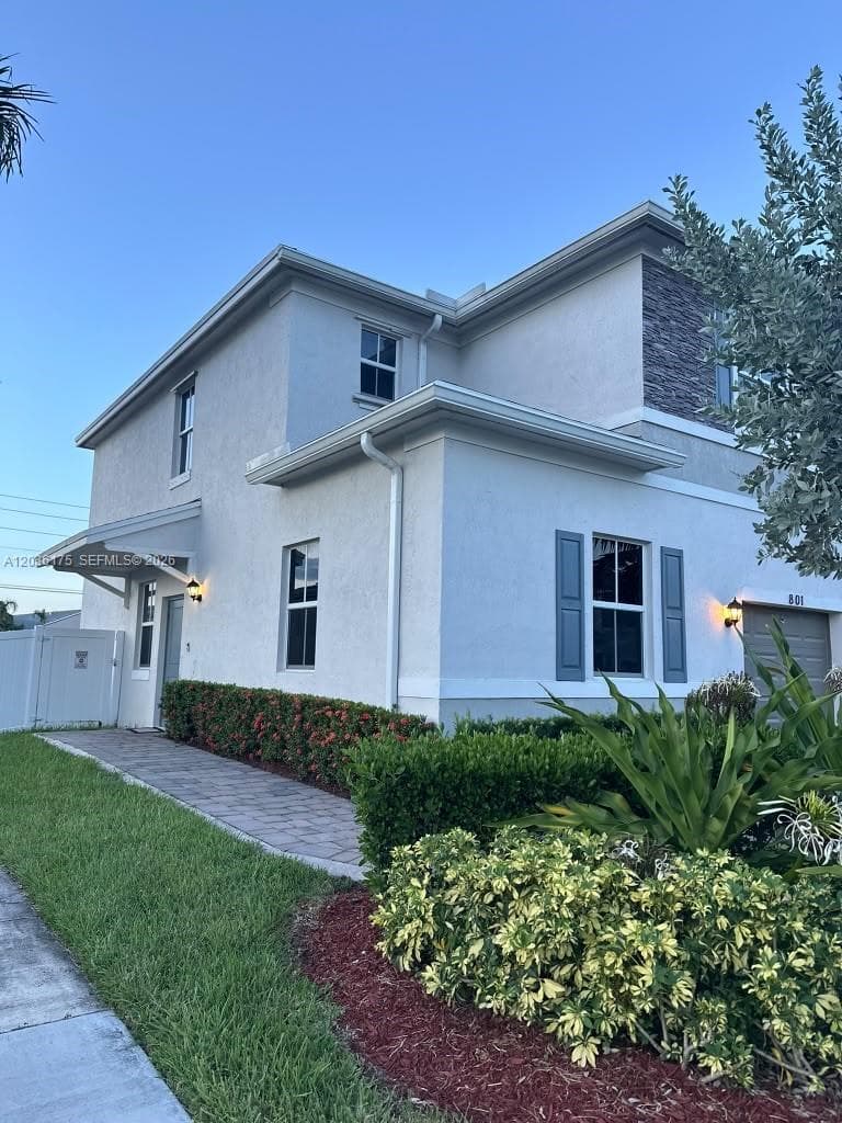 Two-story exterior featuring a light-toned stucco facade, stone veneer accents, decorative shutters, a paver walkway, and mature professional landscaping