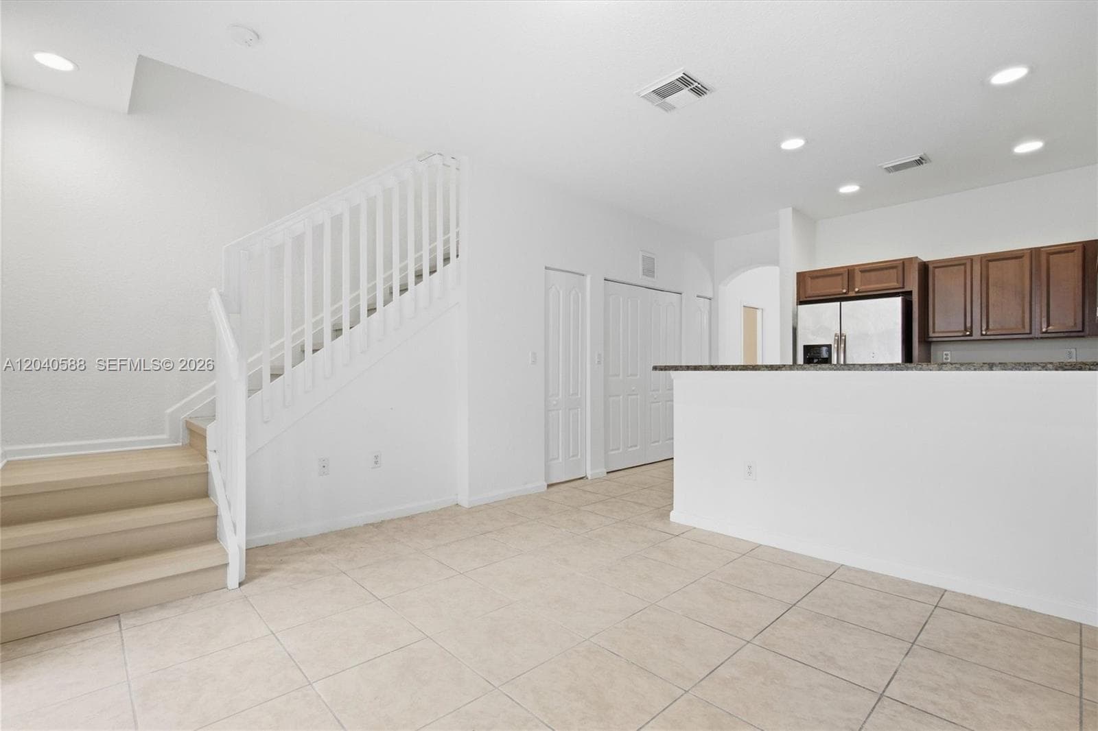 Light-toned tile flooring, recessed ceiling lighting, a staircase with white risers and railing, a kitchen island with a dark countertop, and dark wood-finish cabinetry