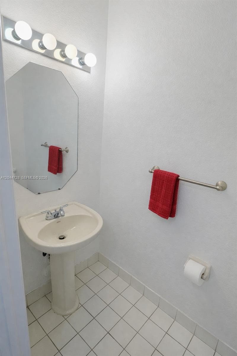 Clean half-bath with pedestal sink, octagonal mirror, bright lighting, and white tile flooring. Simple and well-maintained space.