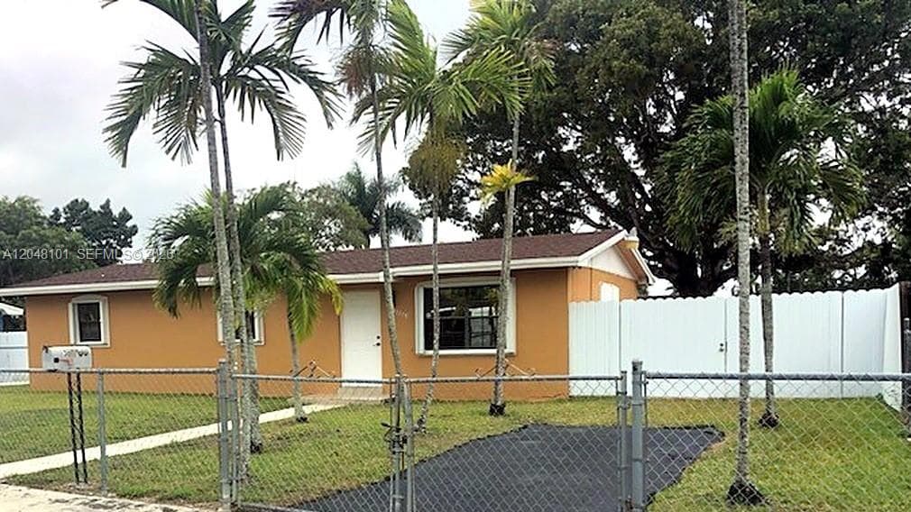 Single-story residence featuring a paved driveway, chain-link perimeter fencing, white vinyl privacy gate, and mature palm trees
