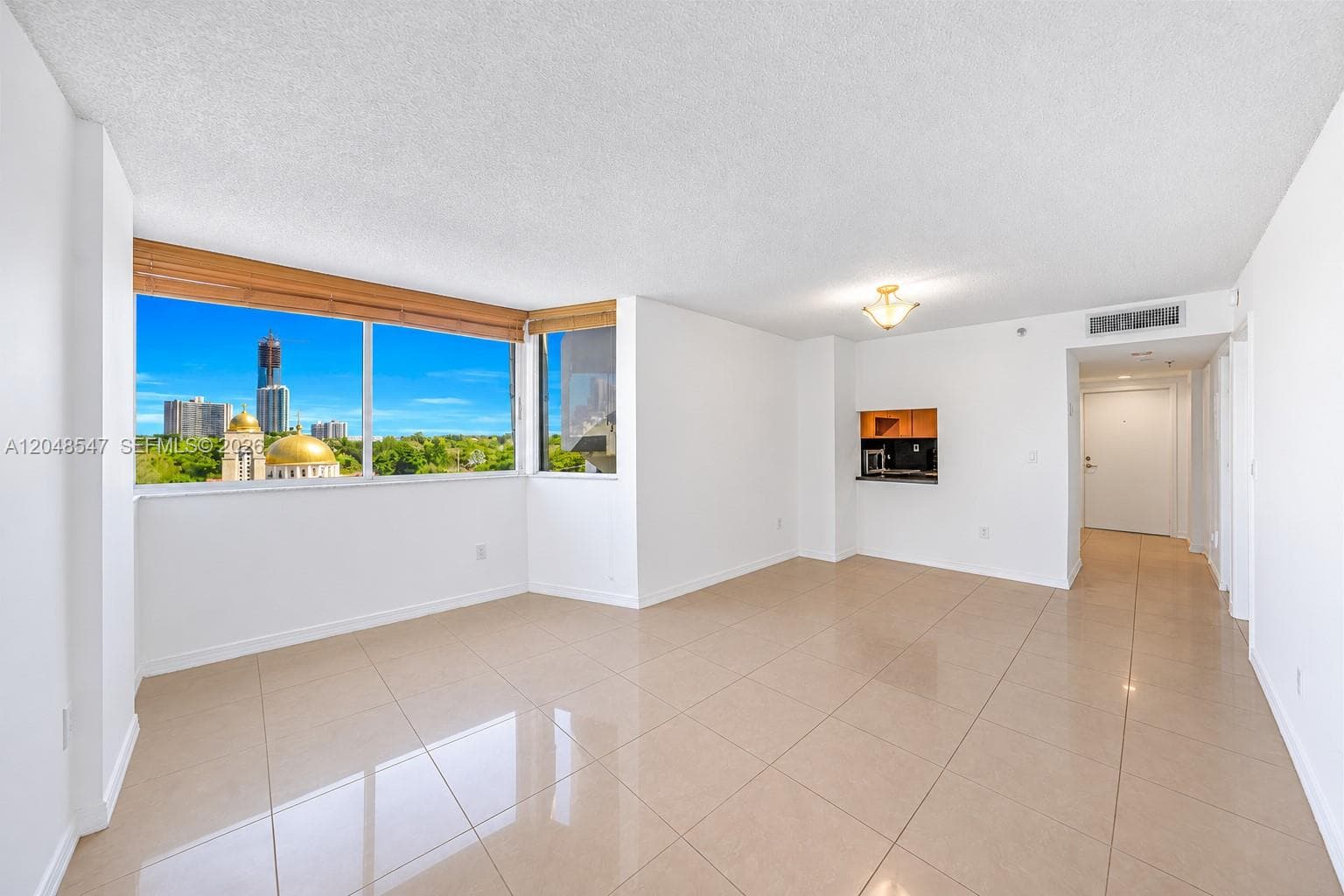 Open-concept interior featuring large-format tile flooring, expansive floor-to-ceiling windows with urban scenery, wood-finish window treatments, and a pass-through kitchen opening