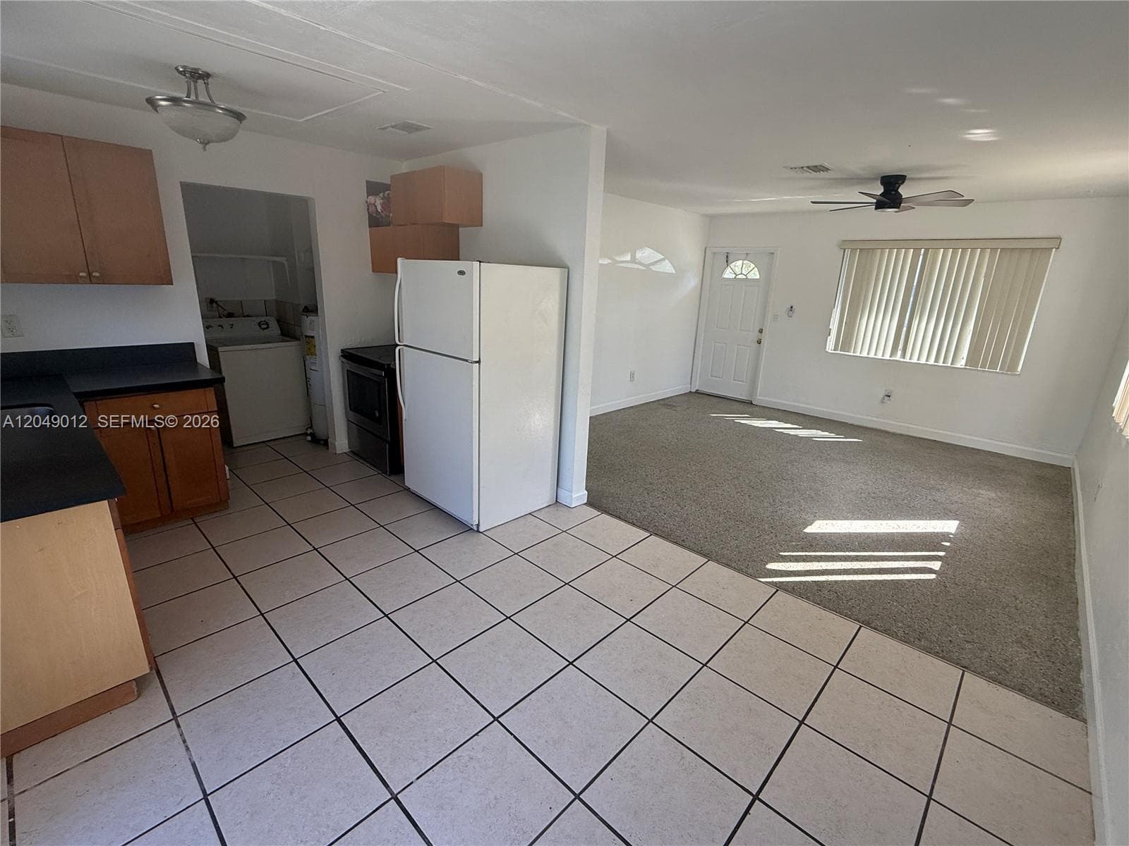 Kitchen featuring tiled flooring, built-in wooden cabinetry, and dark countertops, paired with a carpeted room containing a ceiling fan and vertical window blinds