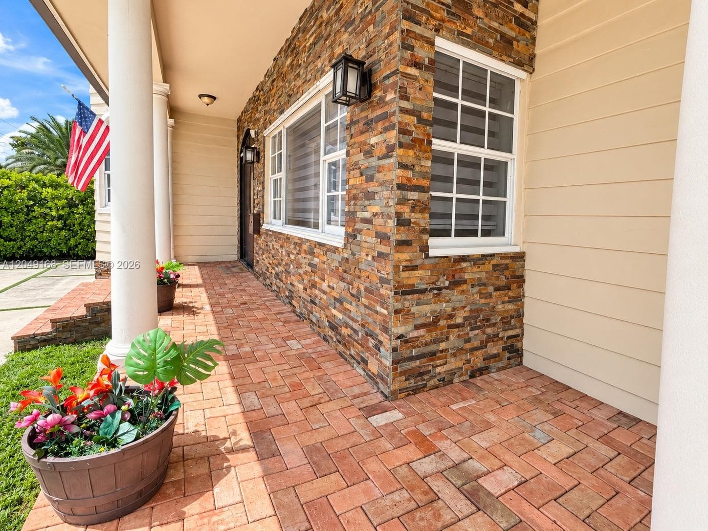 Covered porch featuring herringbone-patterned brick flooring, stone-veneer exterior cladding, horizontal lap siding, structural columns, and wall-mounted exterior lanterns