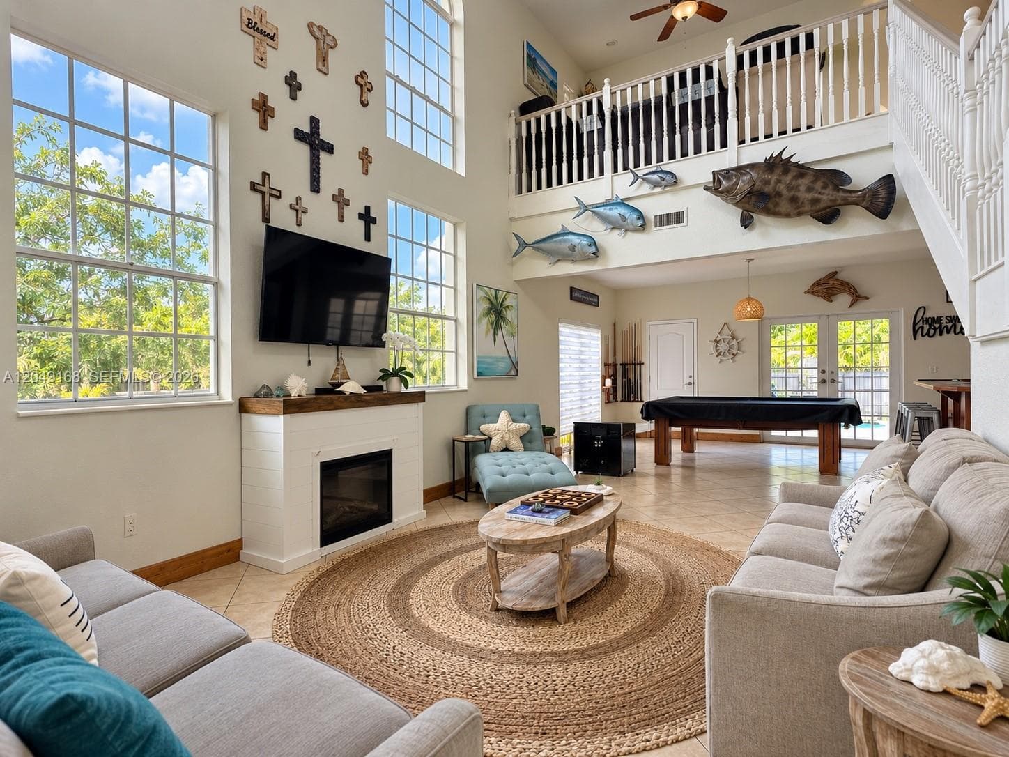 Two-story living area featuring vaulted ceilings, floor-to-ceiling windows, fireplace with a wood mantel, mezzanine overlook with white balustrade, and tile flooring