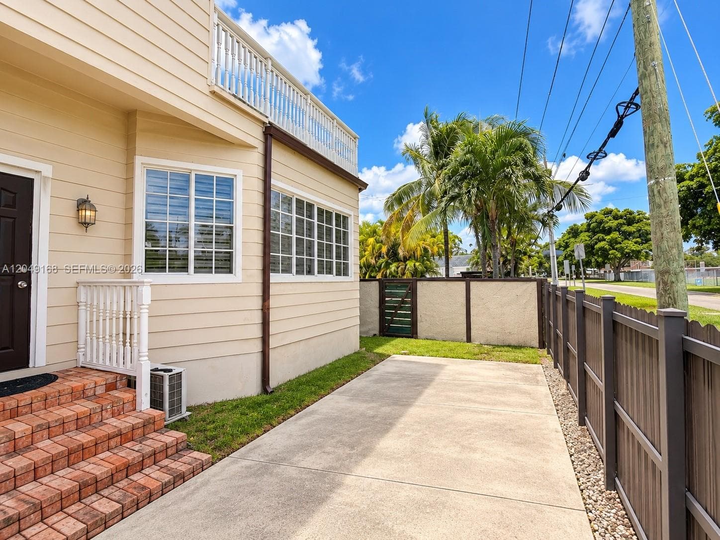 Horizontal lap siding exterior featuring a second-story balcony with white balustrades, brick-paved entry steps, and a private concrete side patio enclosed by wood-style privacy fencing