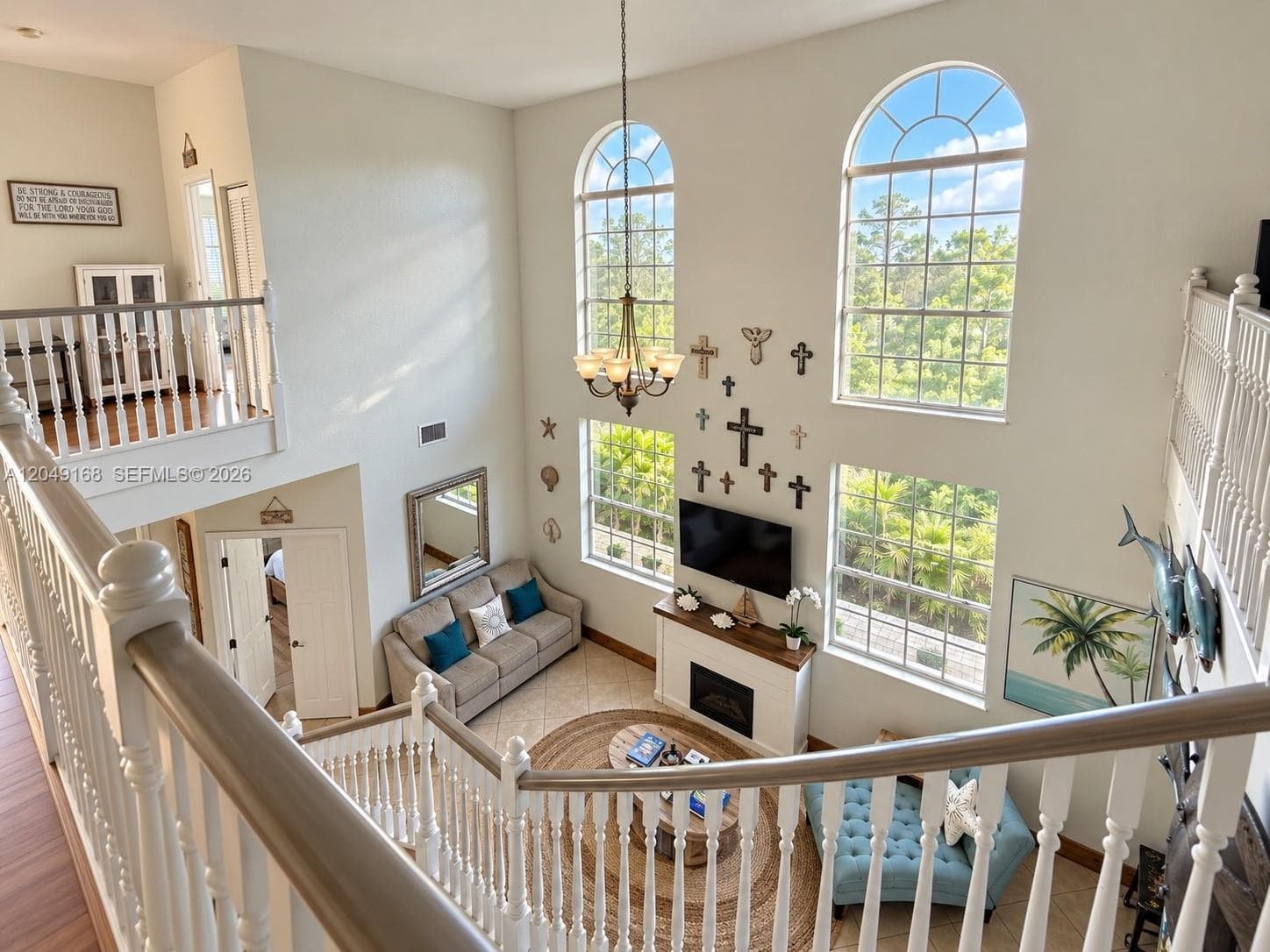 Double-height living area featuring floor-to-ceiling arched windows, a built-in fireplace with a wooden mantel, a tiered chandelier, and a wood-railing staircase with white balusters