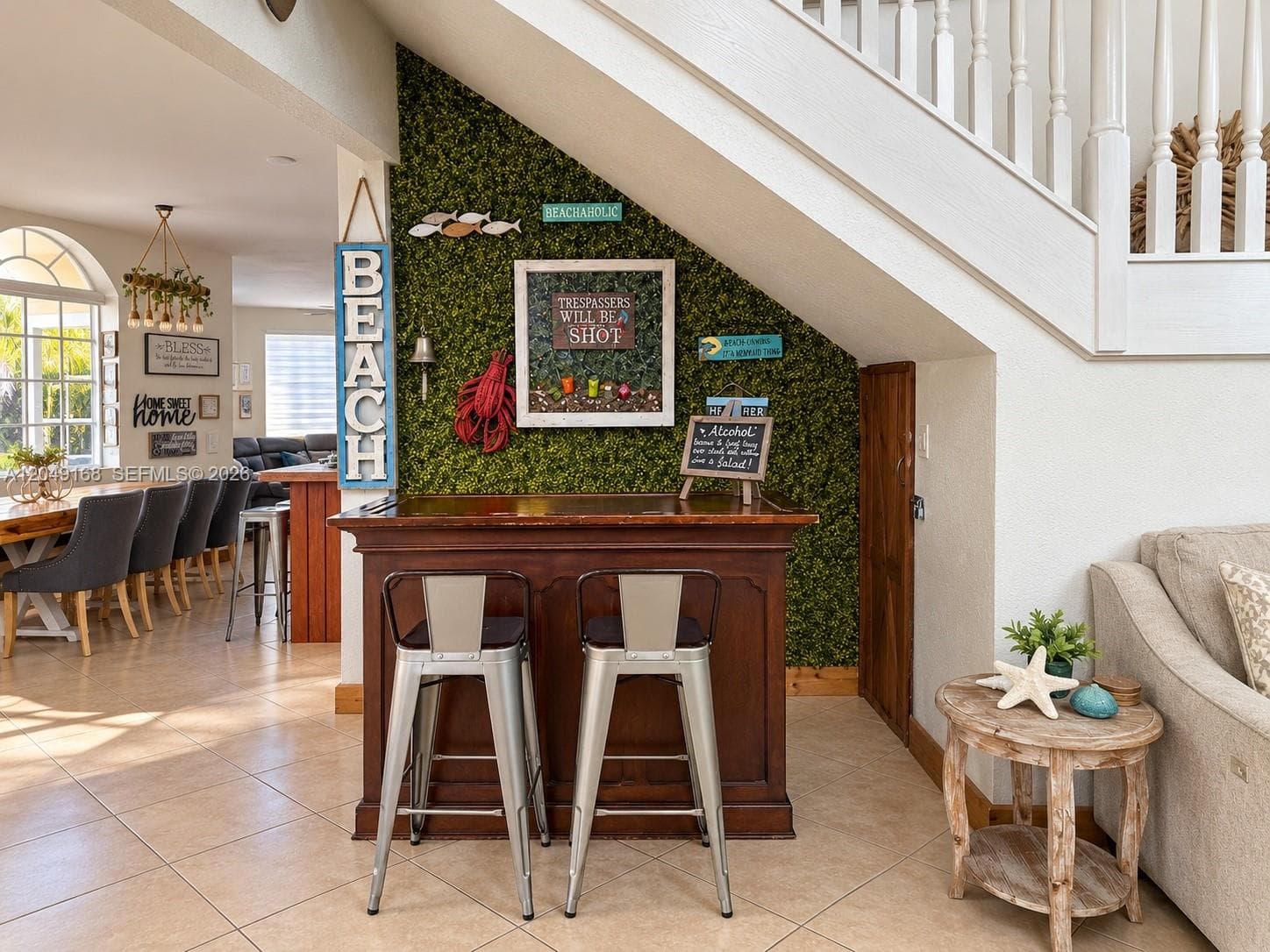 Built-in bar with dark wood cabinetry and a decorative faux-foliage accent wall, complemented by ceramic tile flooring and white wood-railing staircase