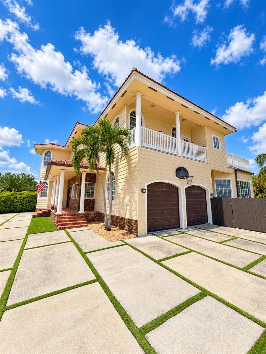 Two-story exterior featuring light-toned siding, stone veneer accents, a second-level balcony with white railings, a covered entryway with column supports, and a segmented concrete driveway with grass inlays