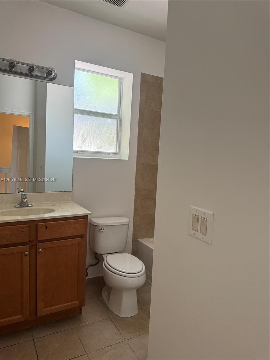 Wood-finish vanity cabinet with a stone-top integrated sink, chrome-finish faucet, wall-mounted mirror with multi-bulb light fixture, and tile flooring