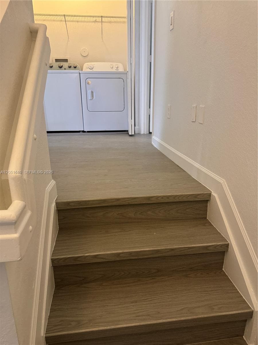 Staircase with wood-finish treads and painted white handrail, leading to a utility area featuring wire shelving and laundry hookups