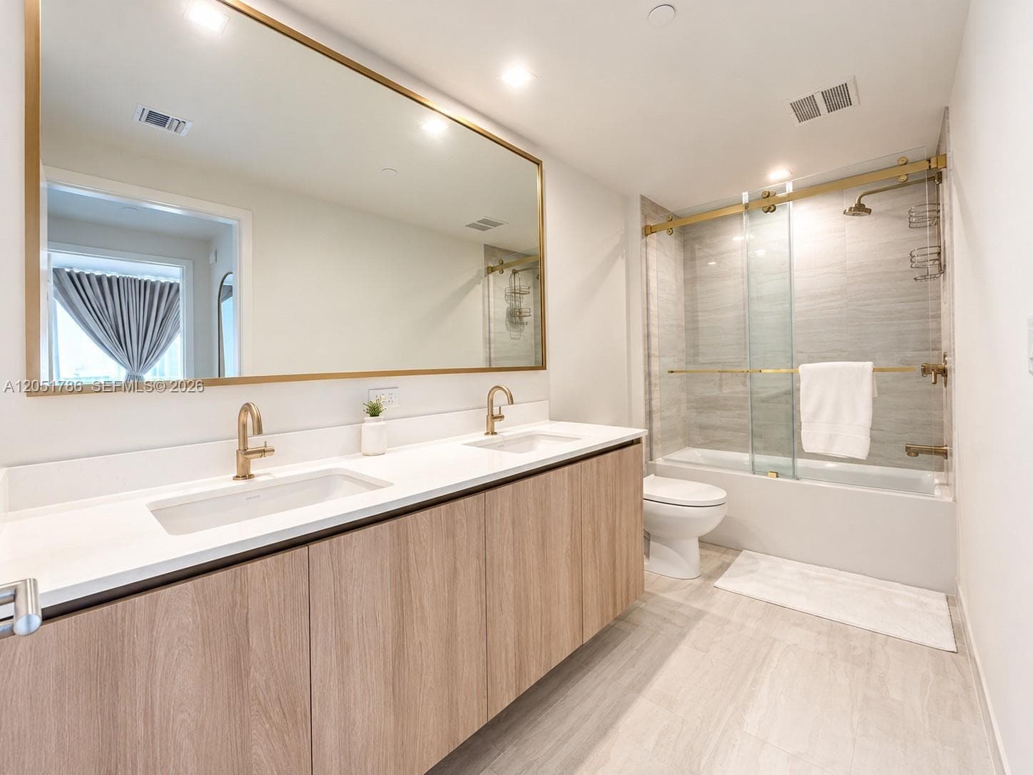 Double vanity featuring a quartz countertop, wood-finish cabinetry, a large brass-framed mirror, and brass-tone fixtures with a matching sliding glass shower enclosure