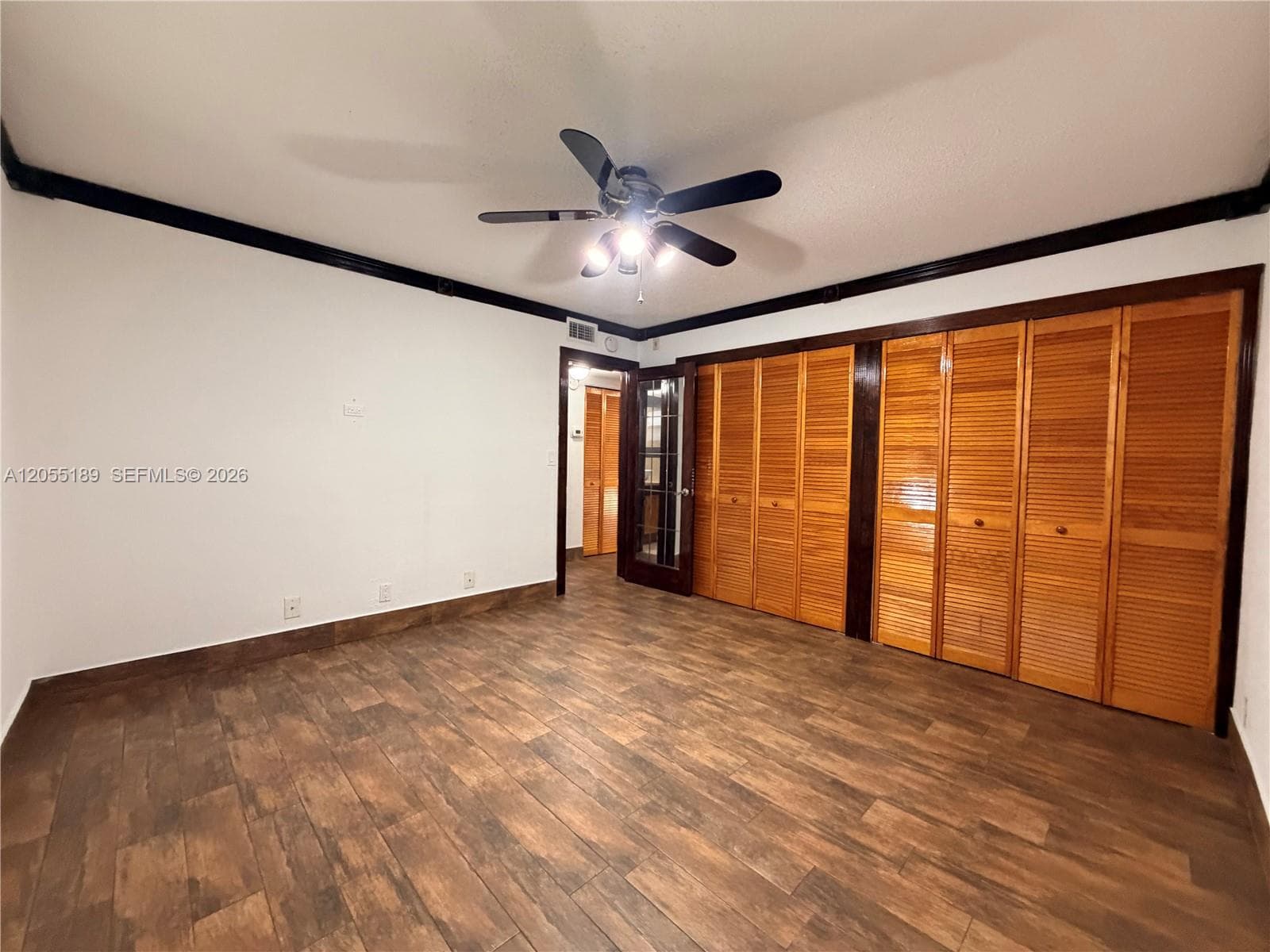 Room featuring wood-finish tile flooring, ceiling fan with integrated lighting, dark-toned crown molding, and extensive louvered-panel built-in closet cabinetry