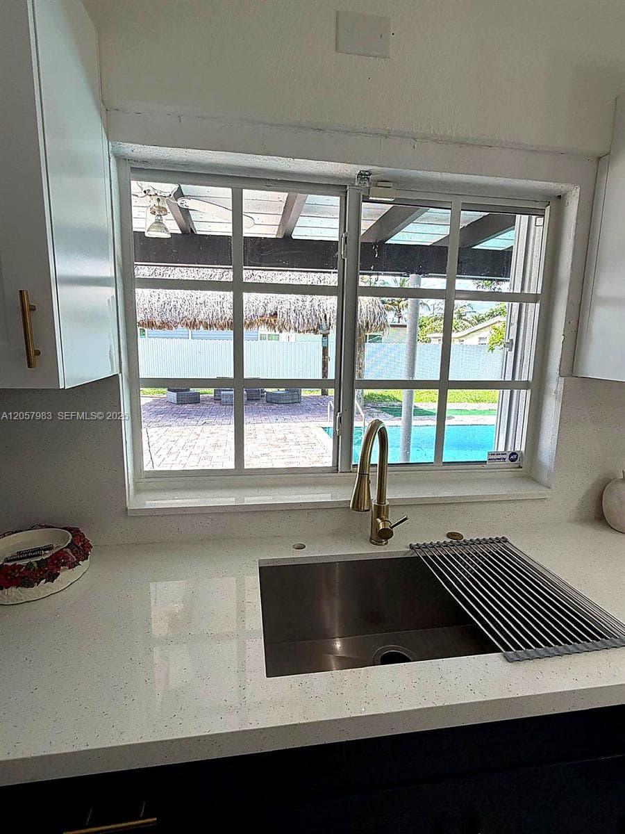 White solid surface countertop, undermount stainless steel sink, gold-finish gooseneck faucet, white upper cabinetry with gold-finish pulls, and a window with a grid pattern