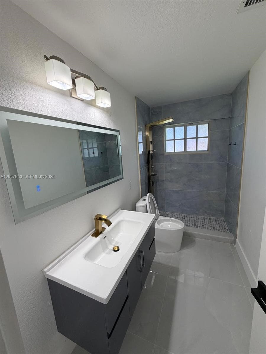 Floating vanity with integrated sink, brass-finish faucet, illuminated mirror, and a walk-in shower with tiled walls