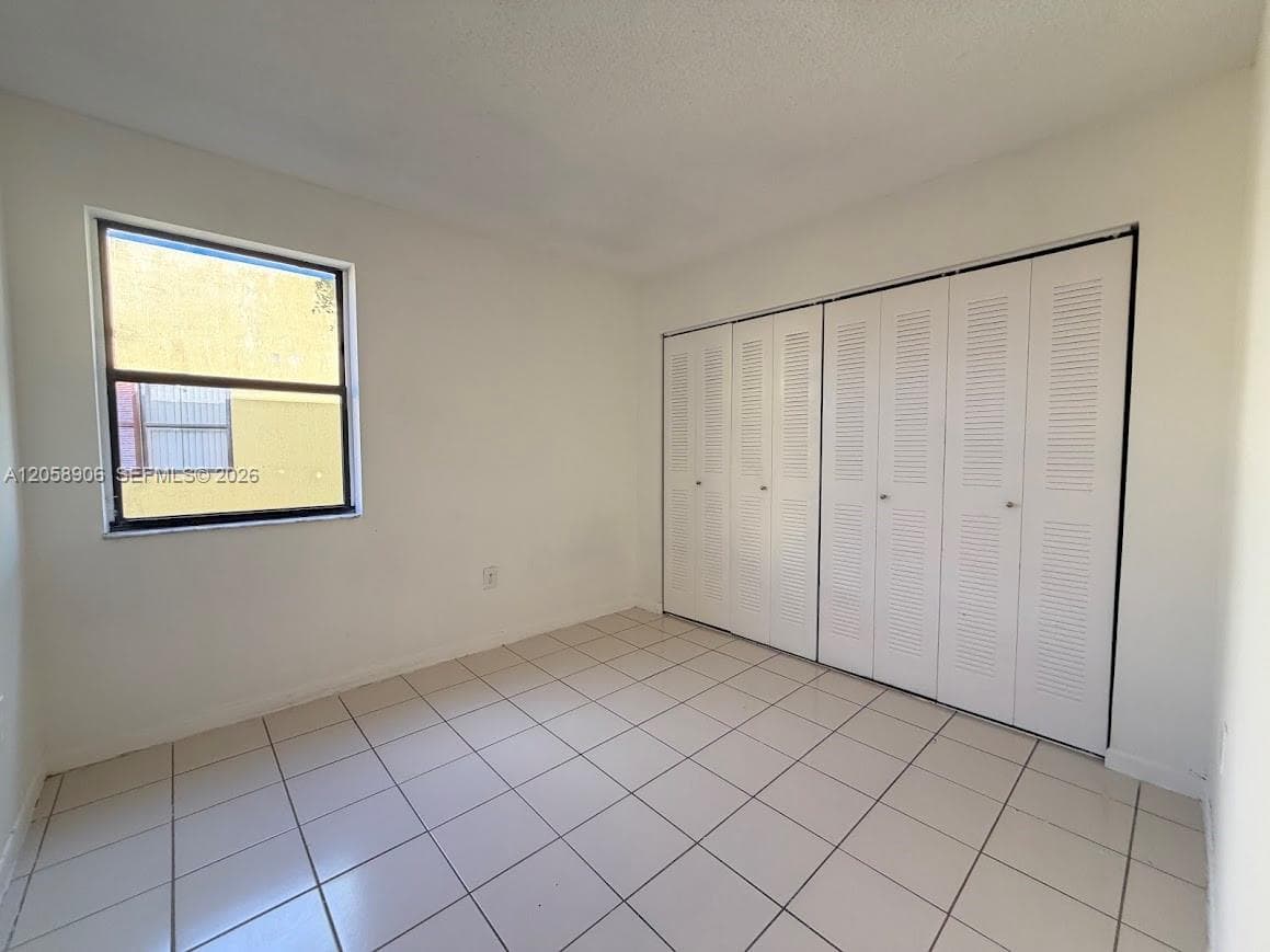 Neutral-toned interior space featuring light-colored square floor tiling, a single-pane window, and floor-to-ceiling bi-fold louvered closet doors